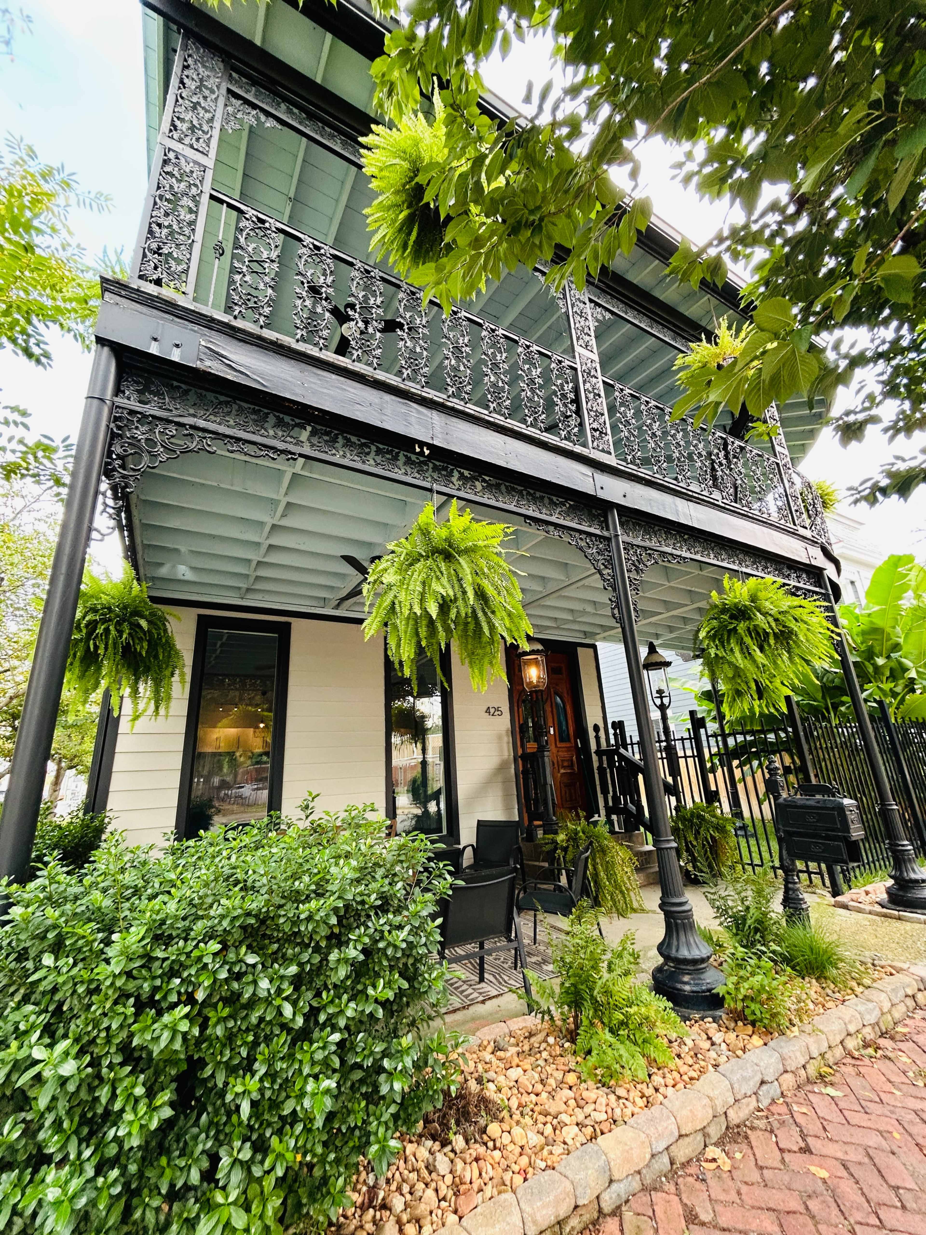 The image shows a two-story house with an ornate, wrought-iron balcony, surrounded by lush greenery and decorative shrubs.