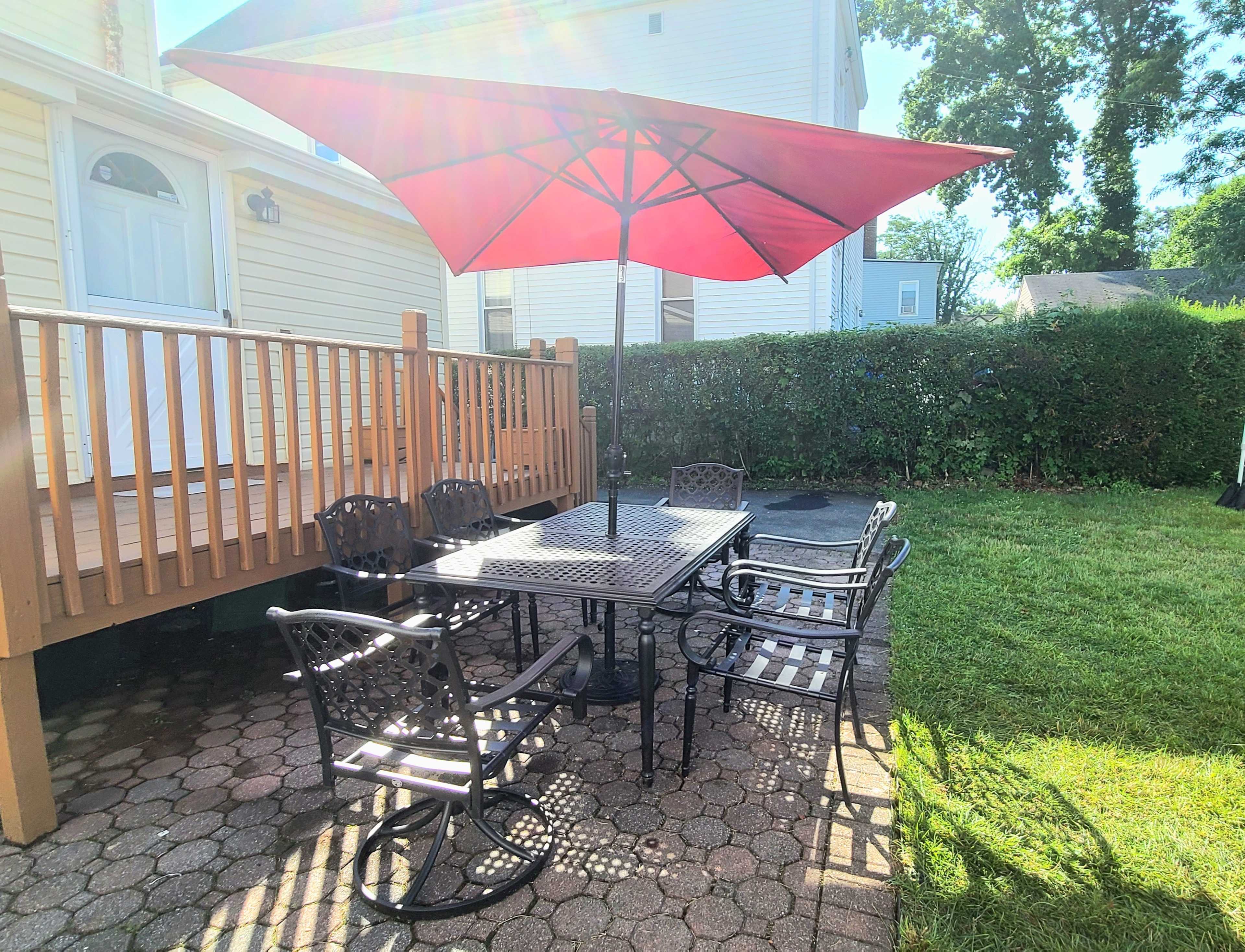 A patio area features a metal table with several chairs beneath a large red umbrella, set on a stone-paved surface next to a grassy lawn.