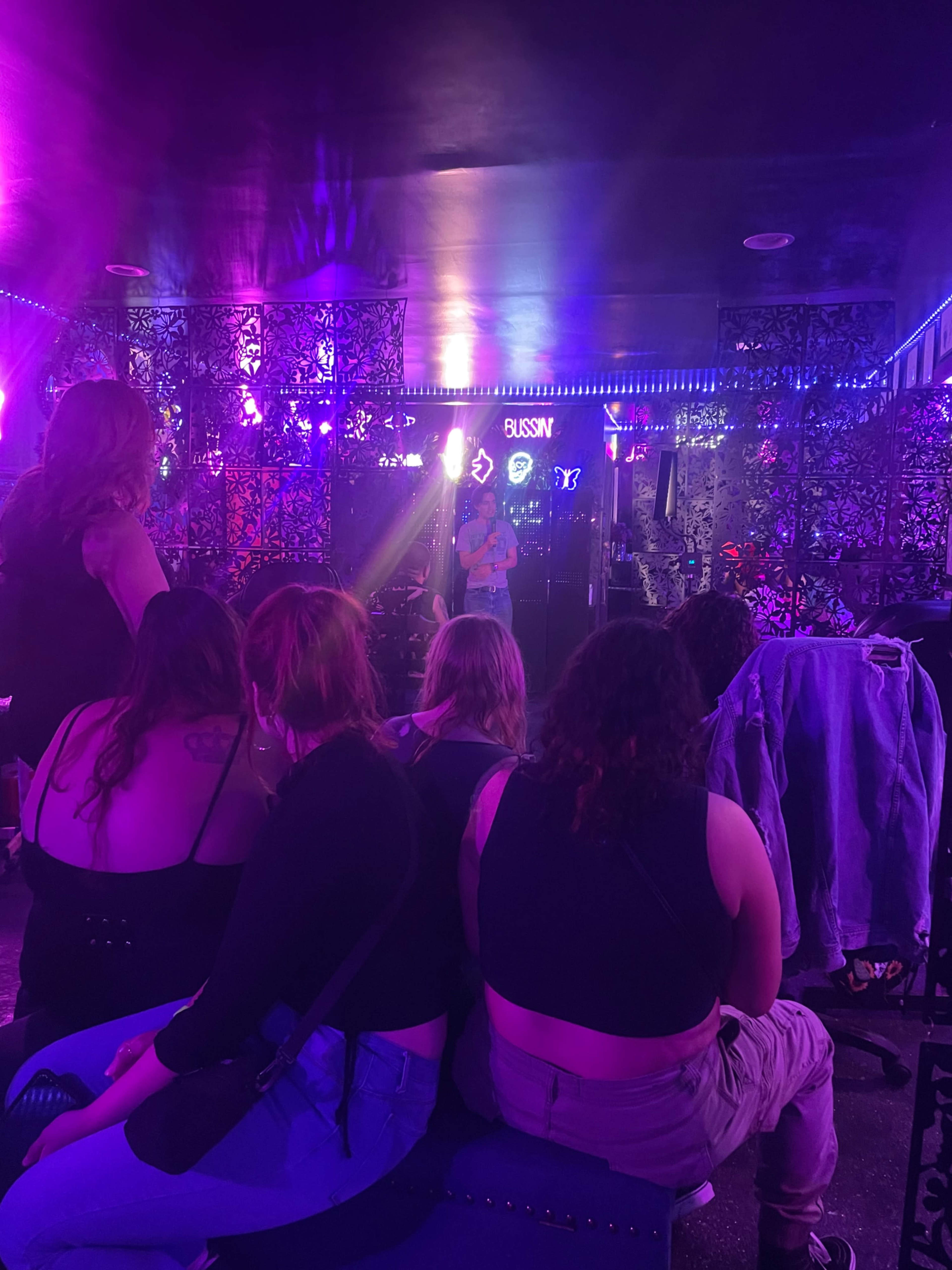 A performer stands on a stage in a dimly lit venue surrounded by patrons seated on couches.