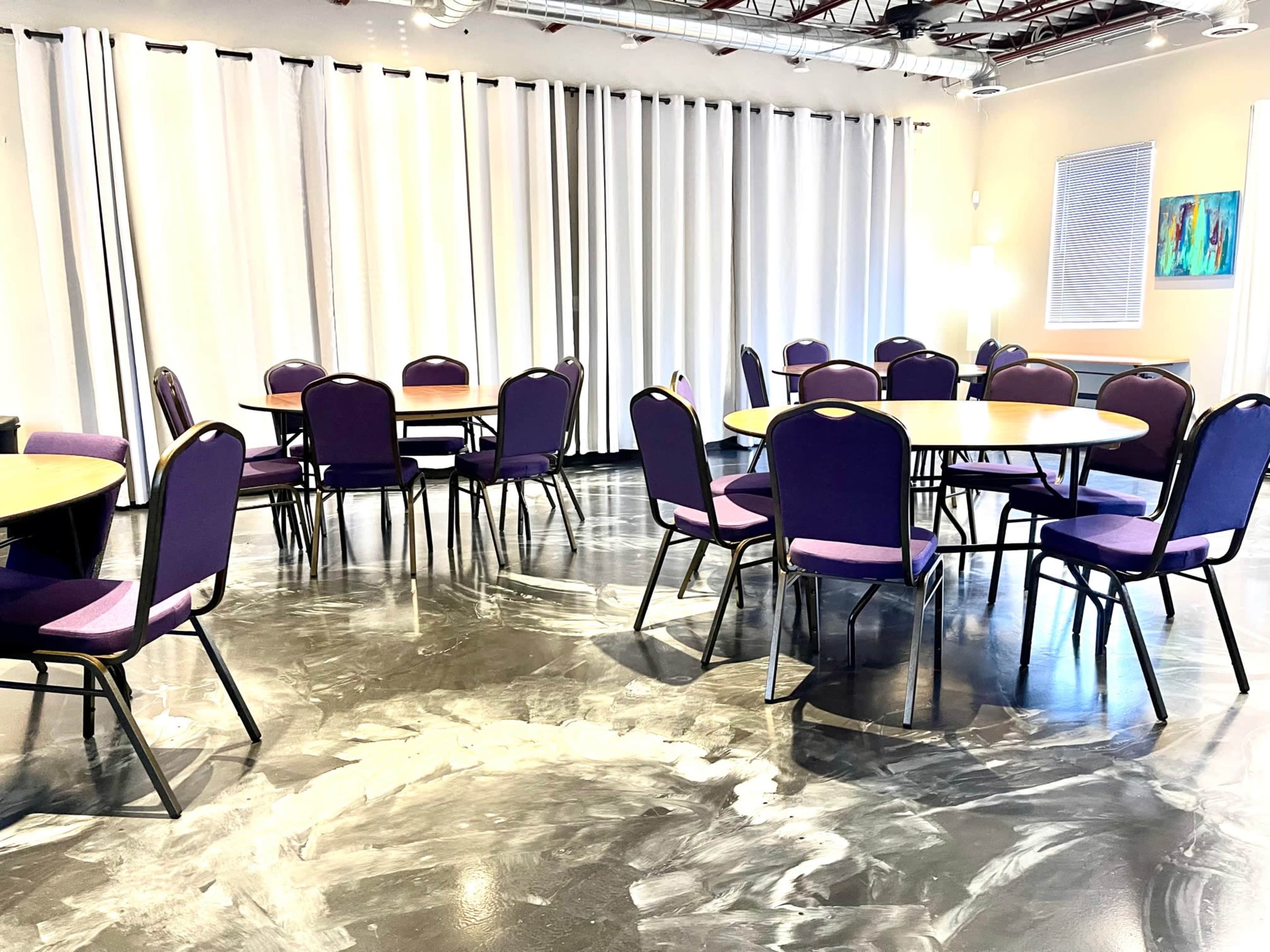 The image shows a room with several round tables surrounded by purple chairs, set against a backdrop of white curtains and windows.