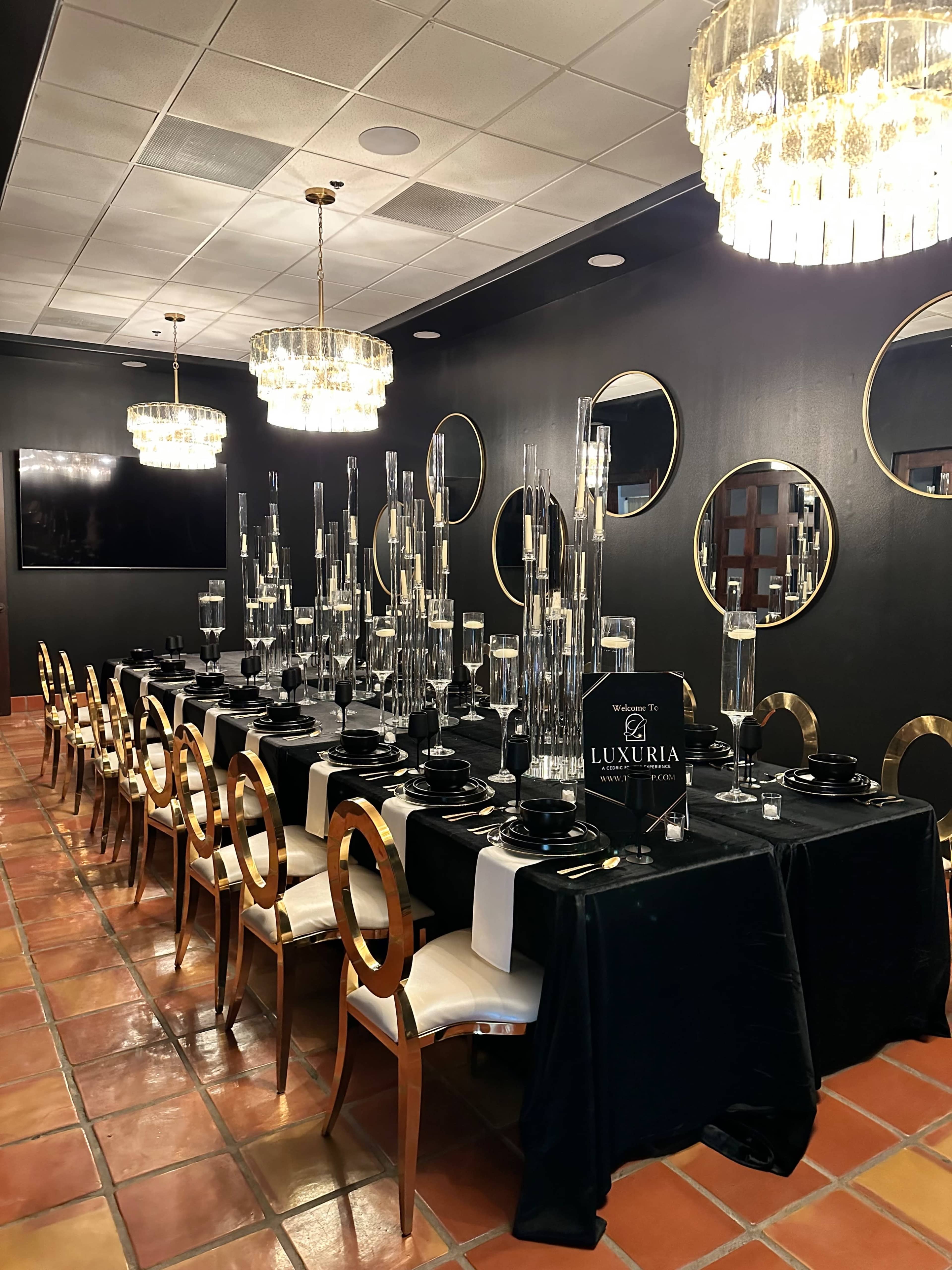 A long dining table is elegantly set in a dark room, featuring tall glass centerpieces and surrounded by gold-accented chairs.