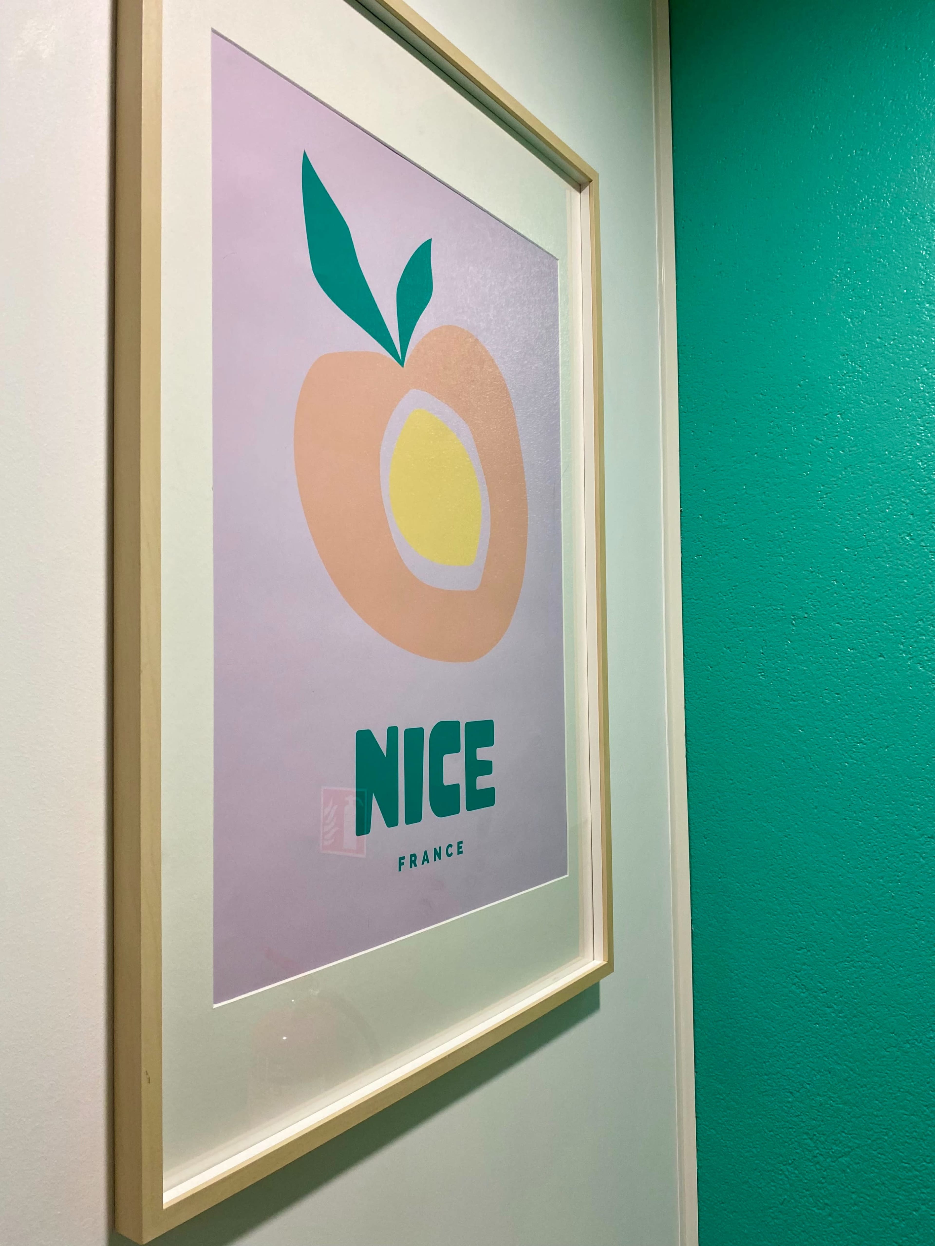 A framed art print featuring a simplified peach illustration with the text "NICE" and "FRANCE" below it, displayed on a wall painted in a turquoise color.