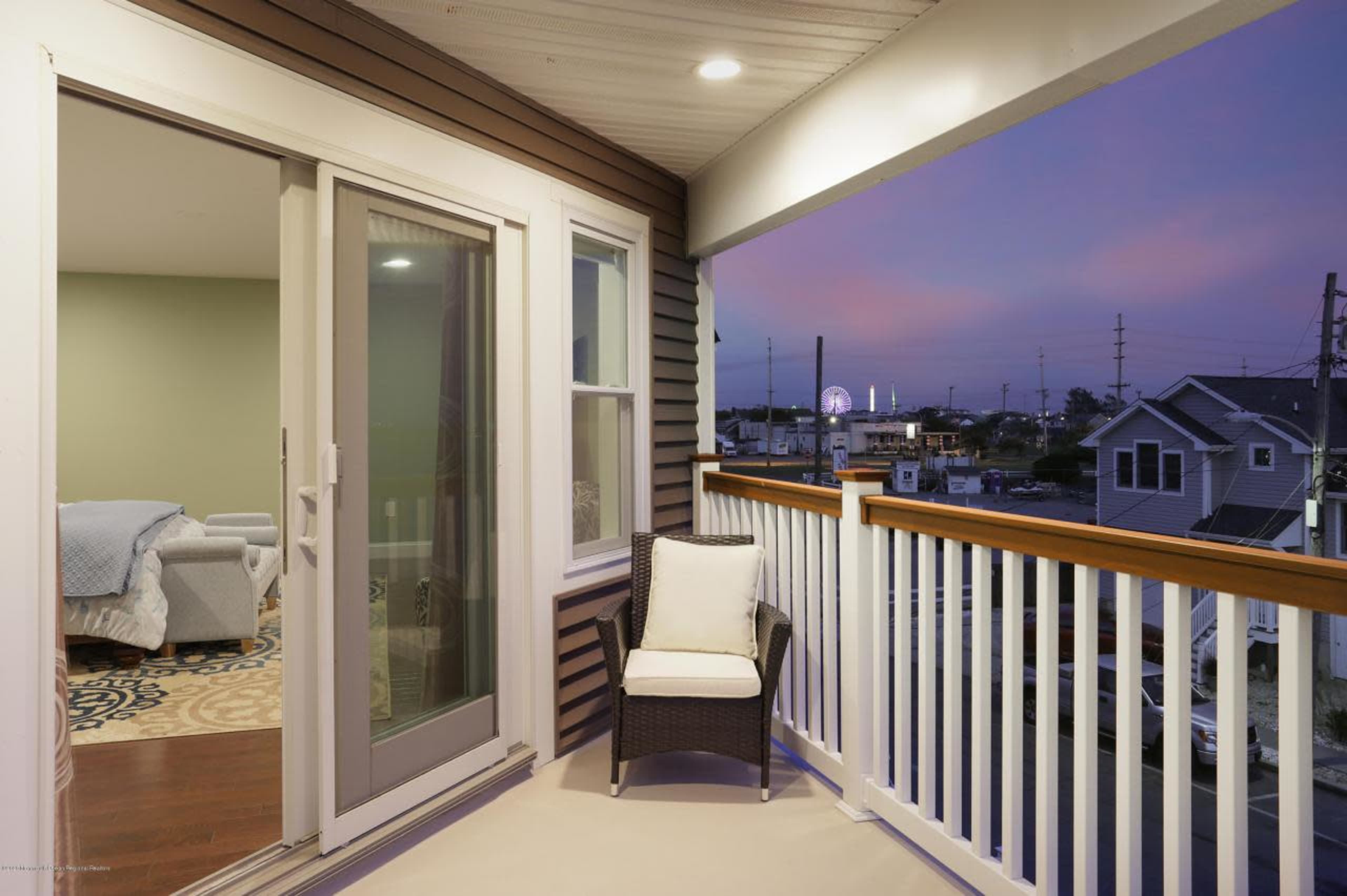 A balcony with a wicker chair overlooking a street and several buildings at dusk.