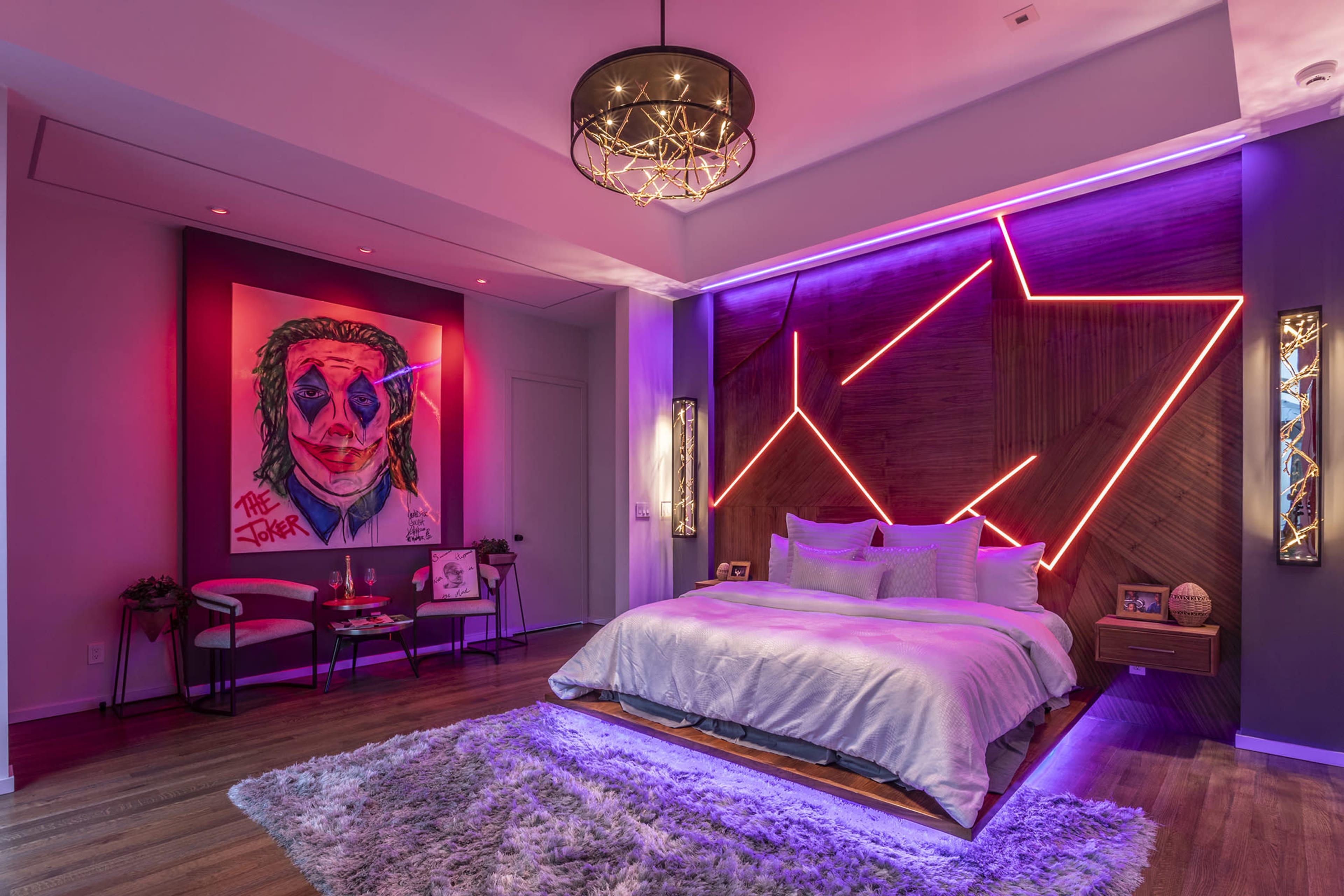 A modern bedroom features a large bed with a light blanket, colorful wall art of the Joker, and neon lighting accenting the walls.