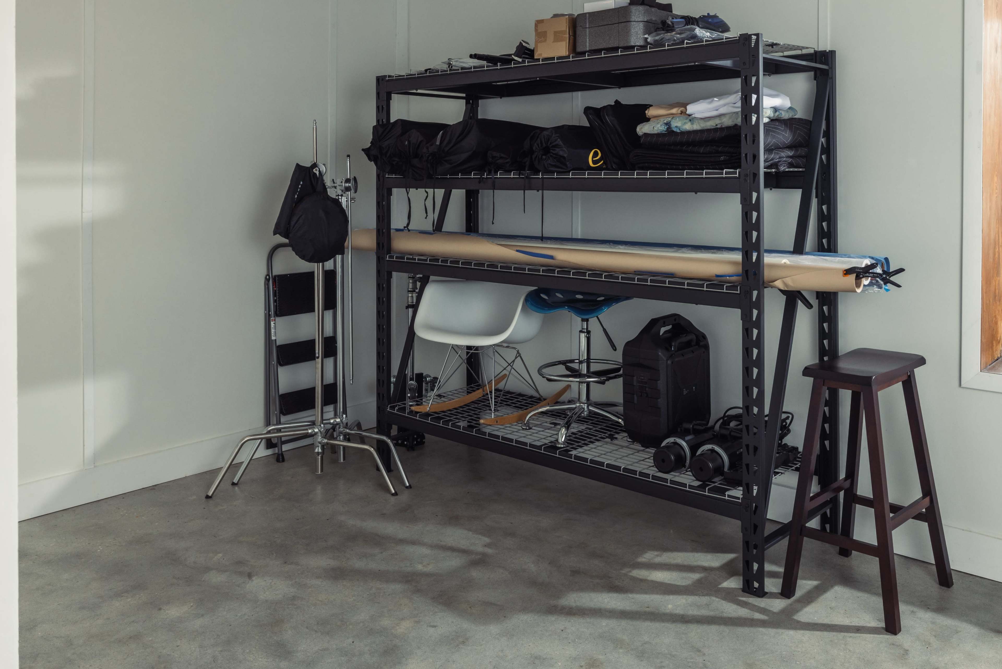 A metal shelving unit displays various items, including clothing, equipment, and a stool, in a corner of a room with concrete flooring.