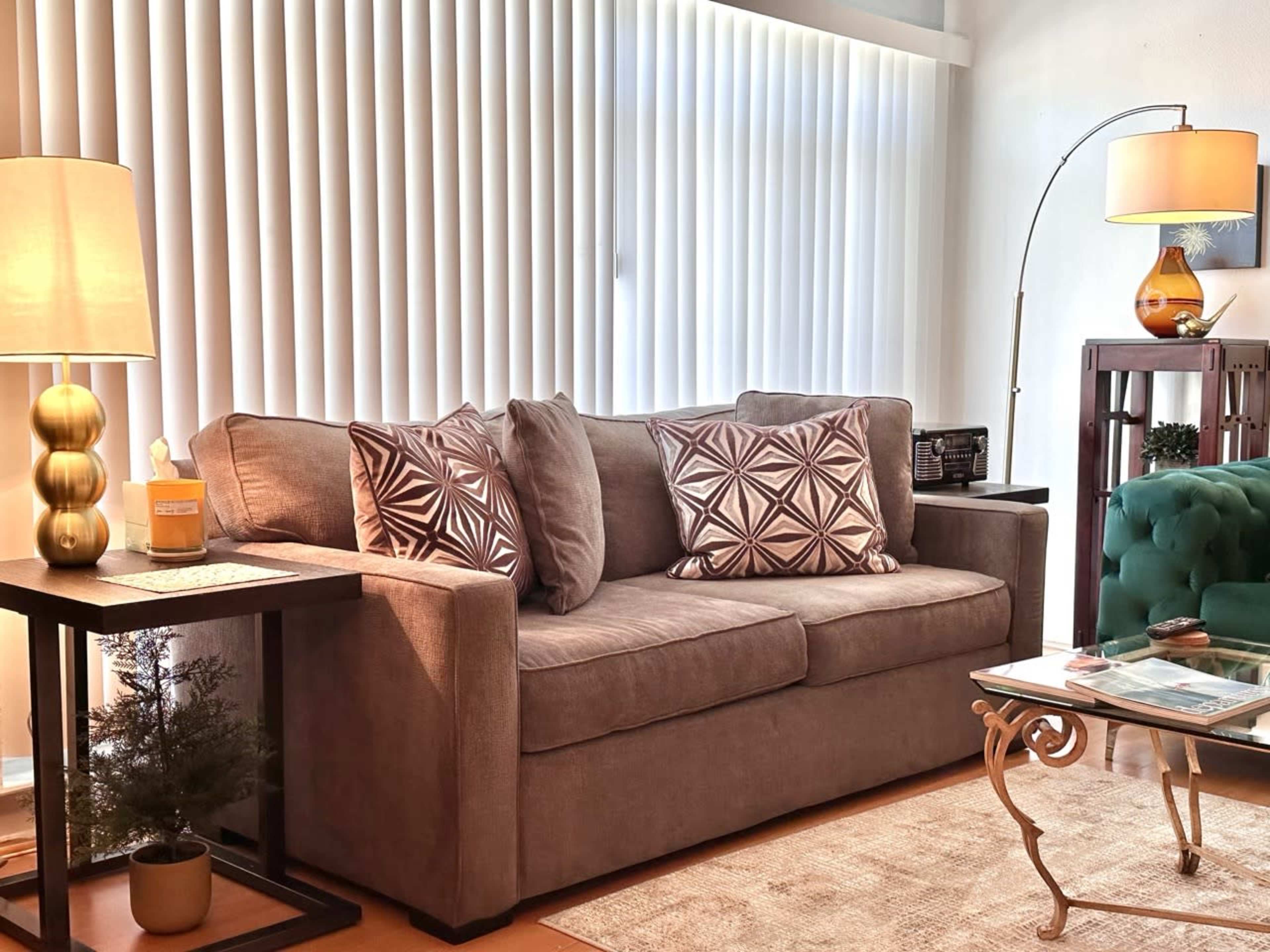The image shows a living room with a brown couch adorned with patterned cushions, a glass coffee table, a floor lamp, and a side table with a decorative object.