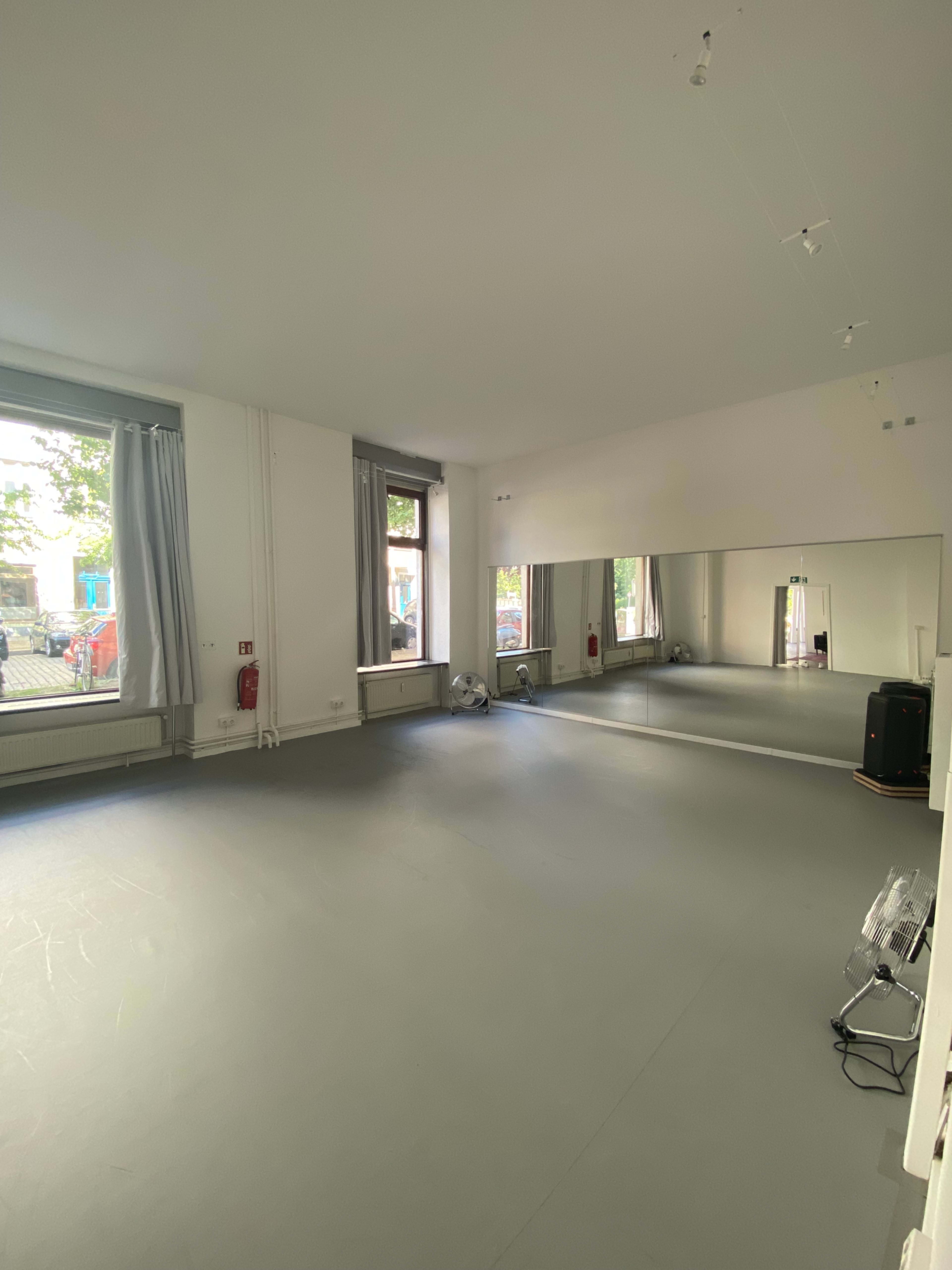 The image shows an empty dance studio with wooden floors, large mirrors on one wall, and windows allowing natural light to enter the space.