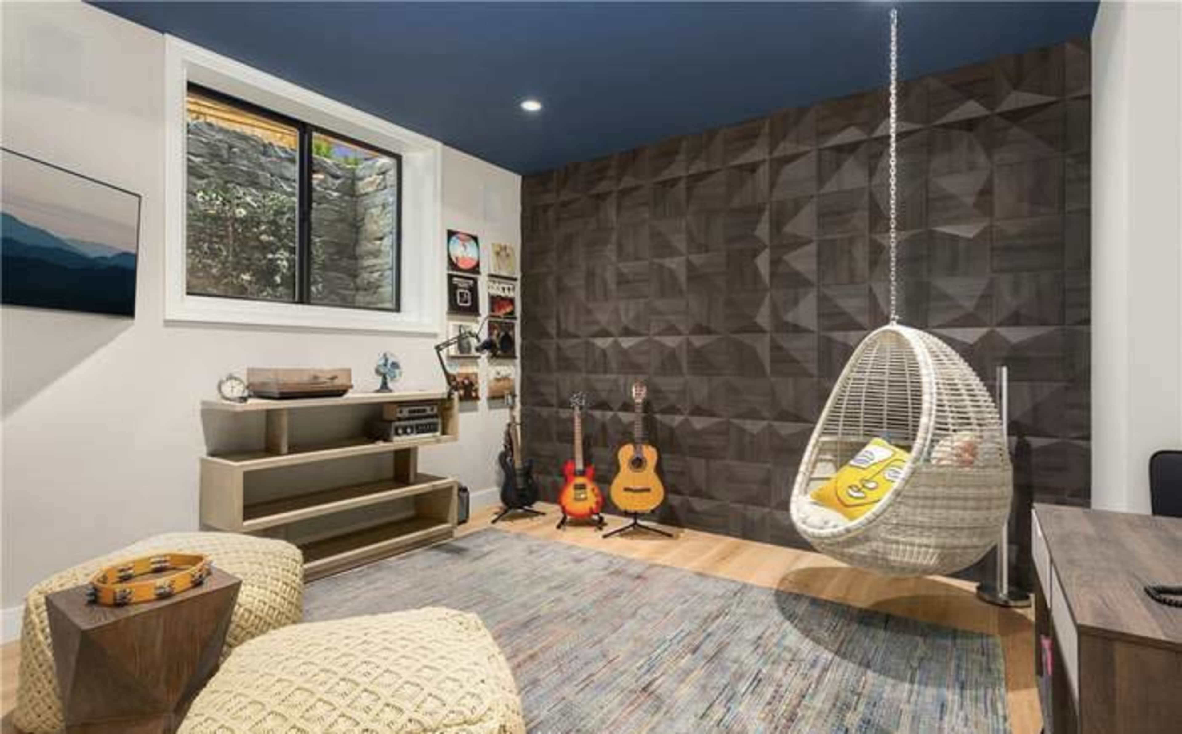 A modern room featuring a hanging chair, two guitars, and a geometric accent wall, with large windows letting in natural light.