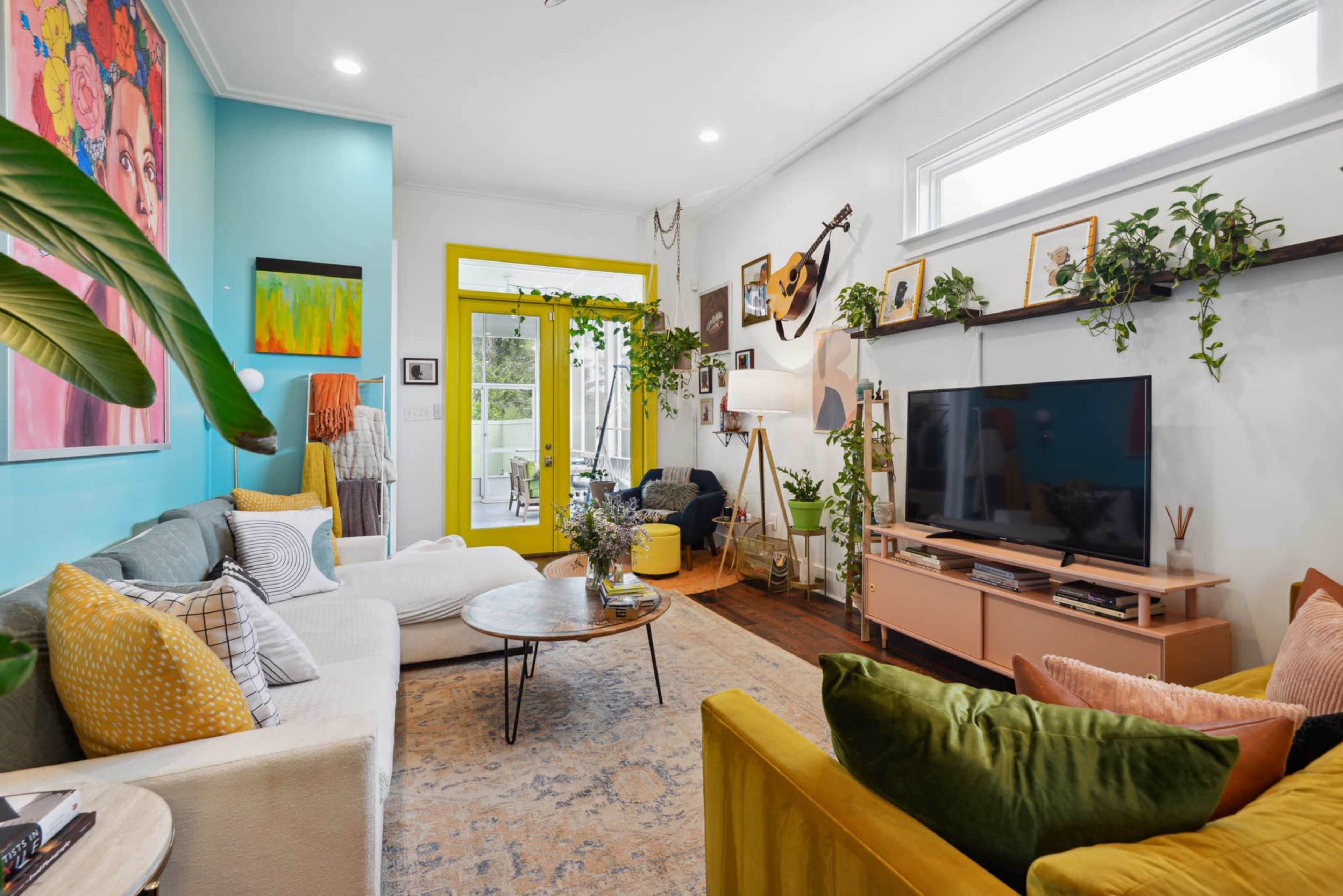 A colorful living room features two sofas, a round coffee table, a television on a light wood console, and plants decorating the walls and furniture.