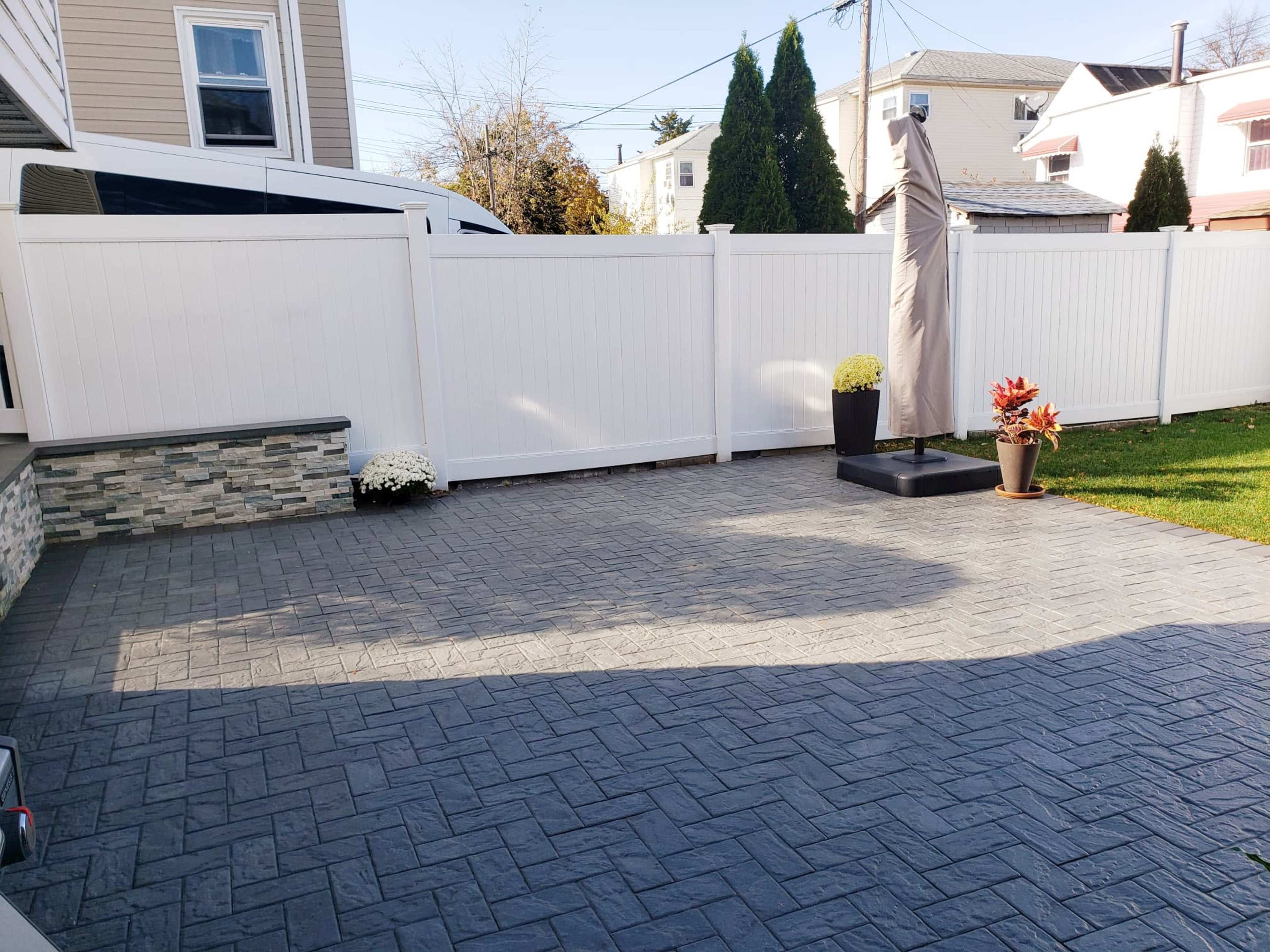 The image shows a patio area with textured concrete flooring, a raised stone border, potted plants, and a large umbrella stand against a white fence.