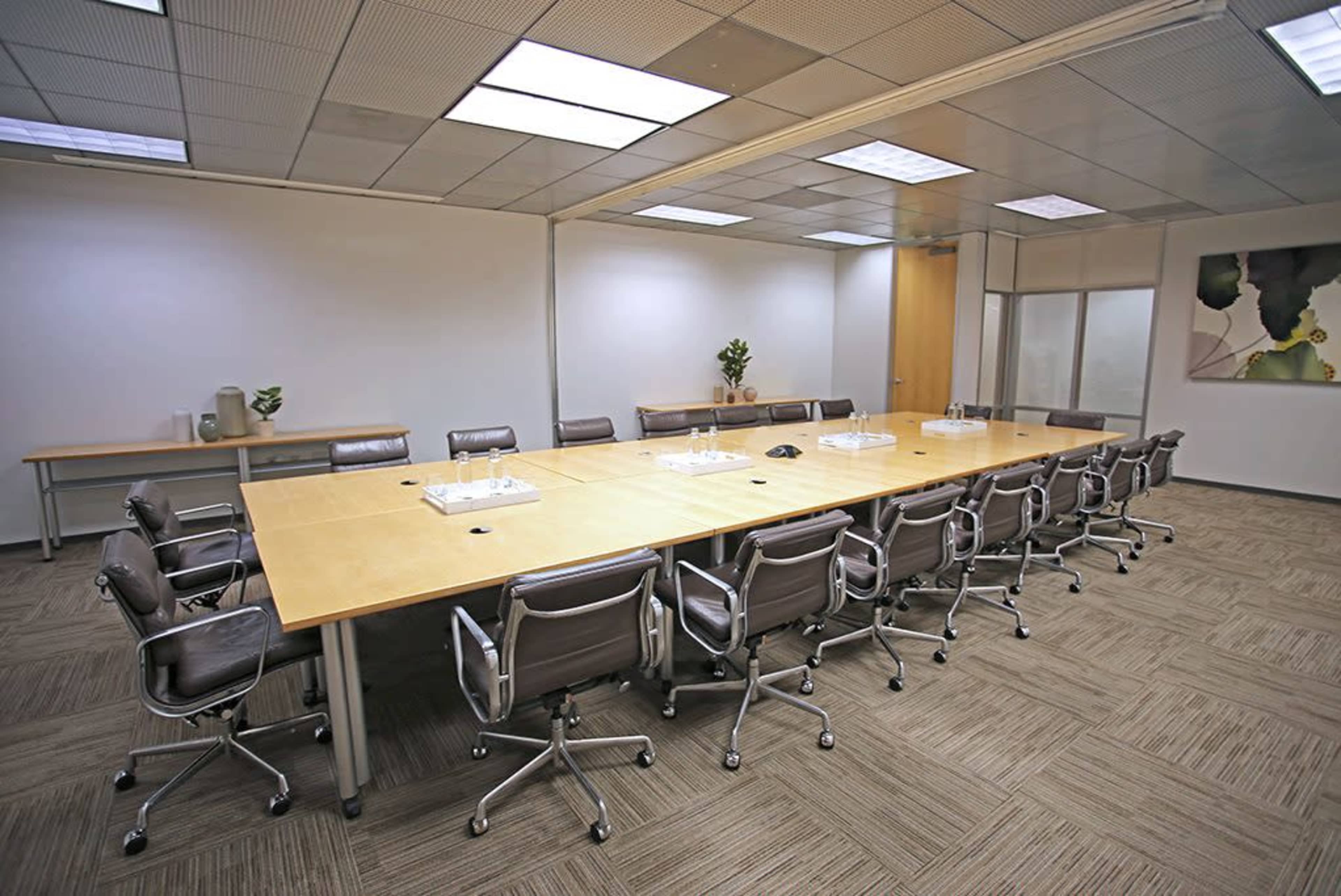 A spacious conference room with a large wooden table surrounded by several gray office chairs, and a plant in a corner.