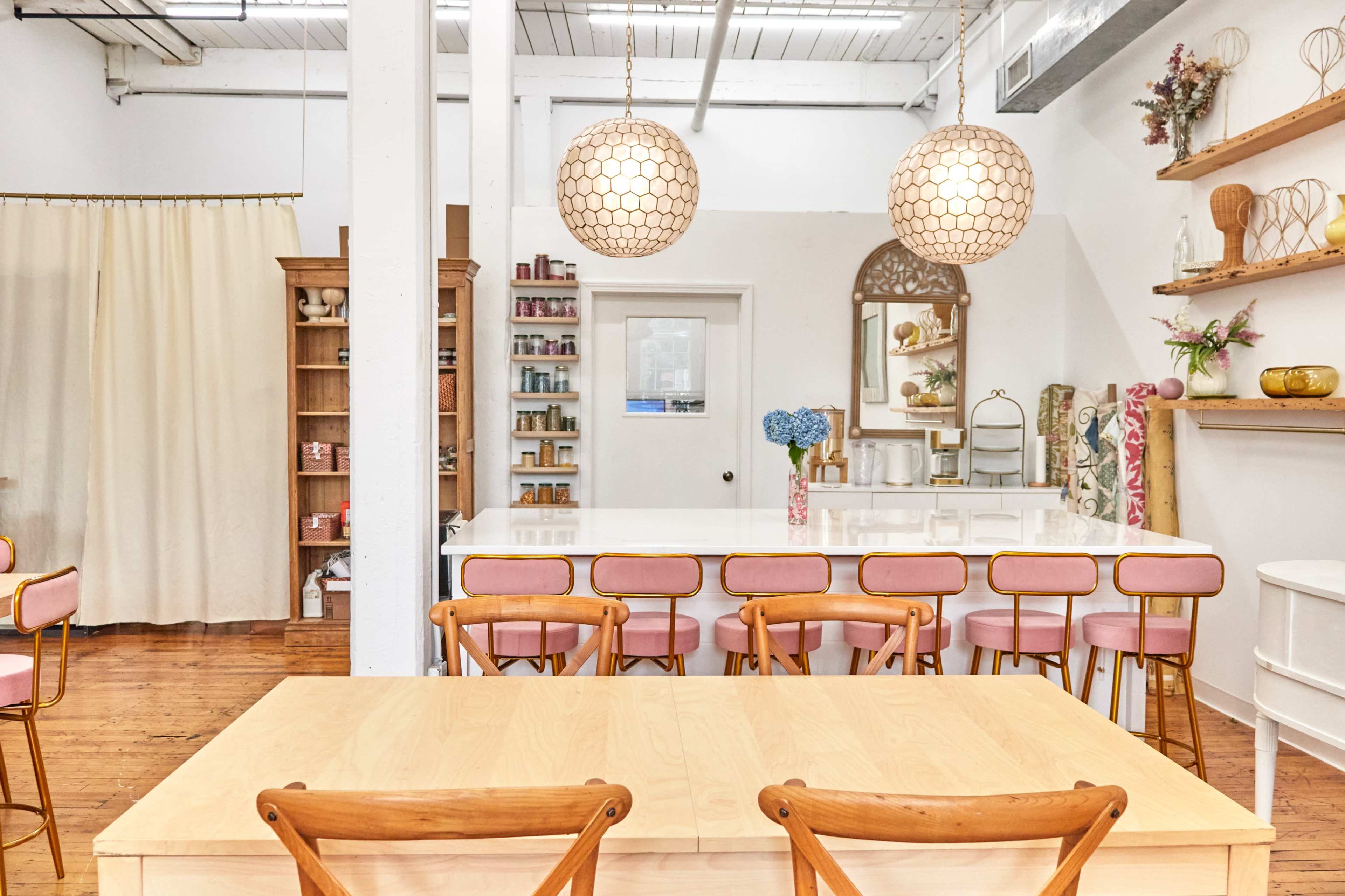 A bright interior with a long white counter, pink upholstered stools, a wooden dining table, and decorative shelves against the walls.