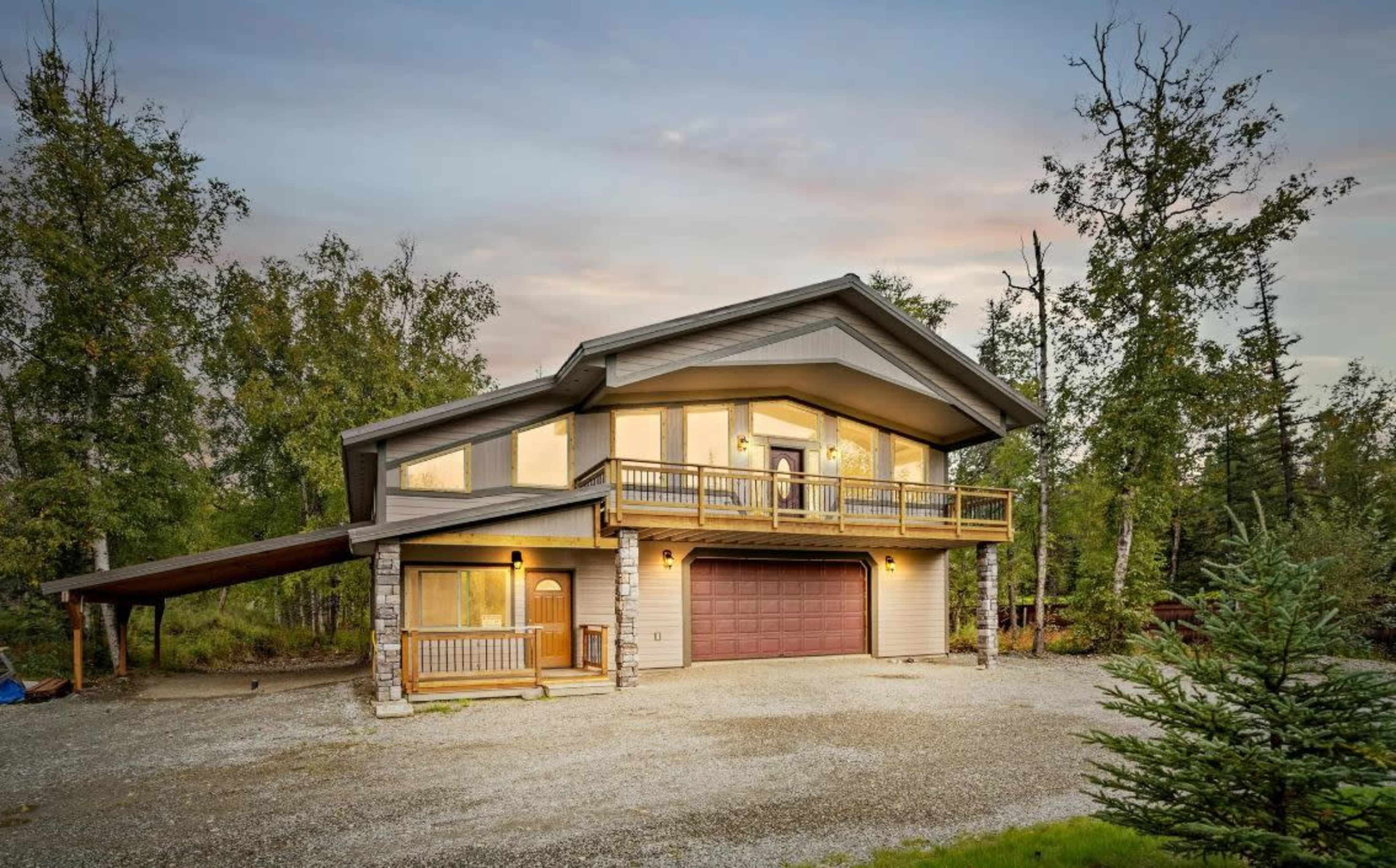 A modern two-story house with a large front deck and a garage is surrounded by trees and gravel driveway.