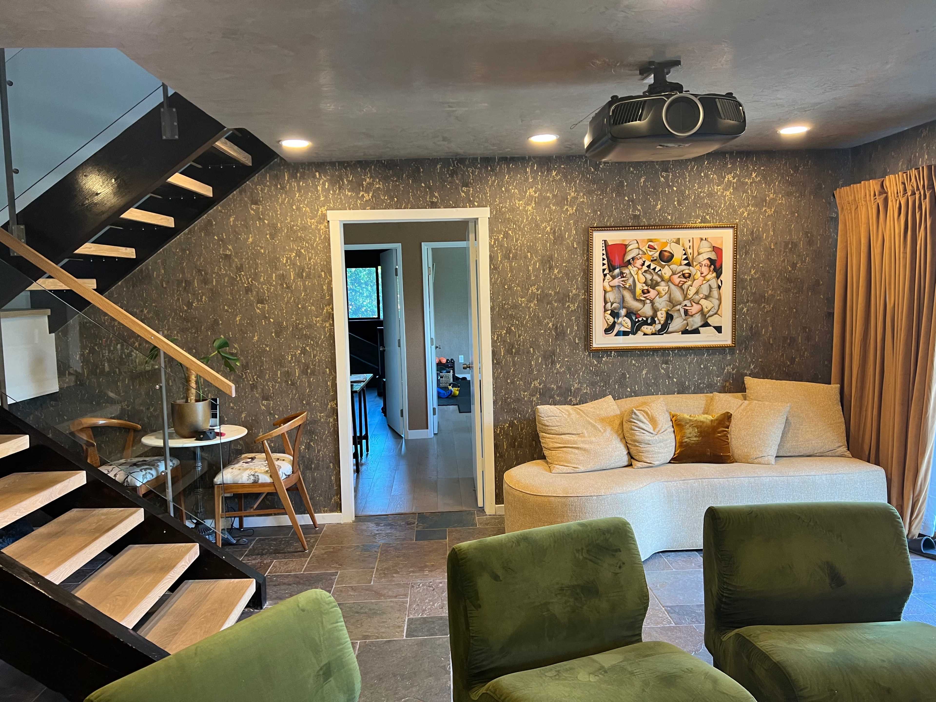 The image shows a modern living area with a couch, two green chairs, a staircase, a table with two chairs, and a projector mounted on the ceiling, along with artwork on the wall.