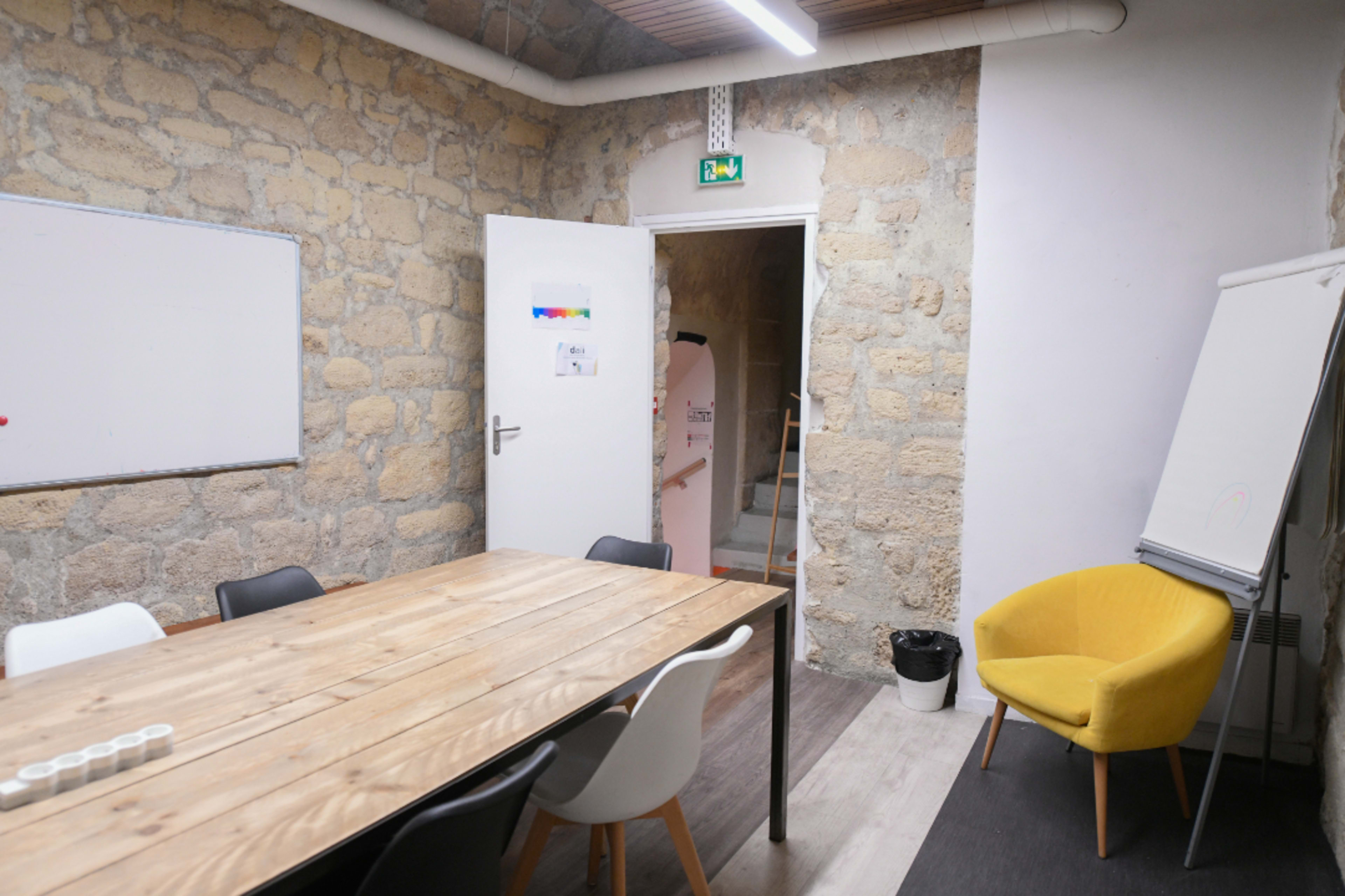A meeting room with a wooden table and chairs, a yellow armchair in the corner, and a doorway leading to a staircase.
