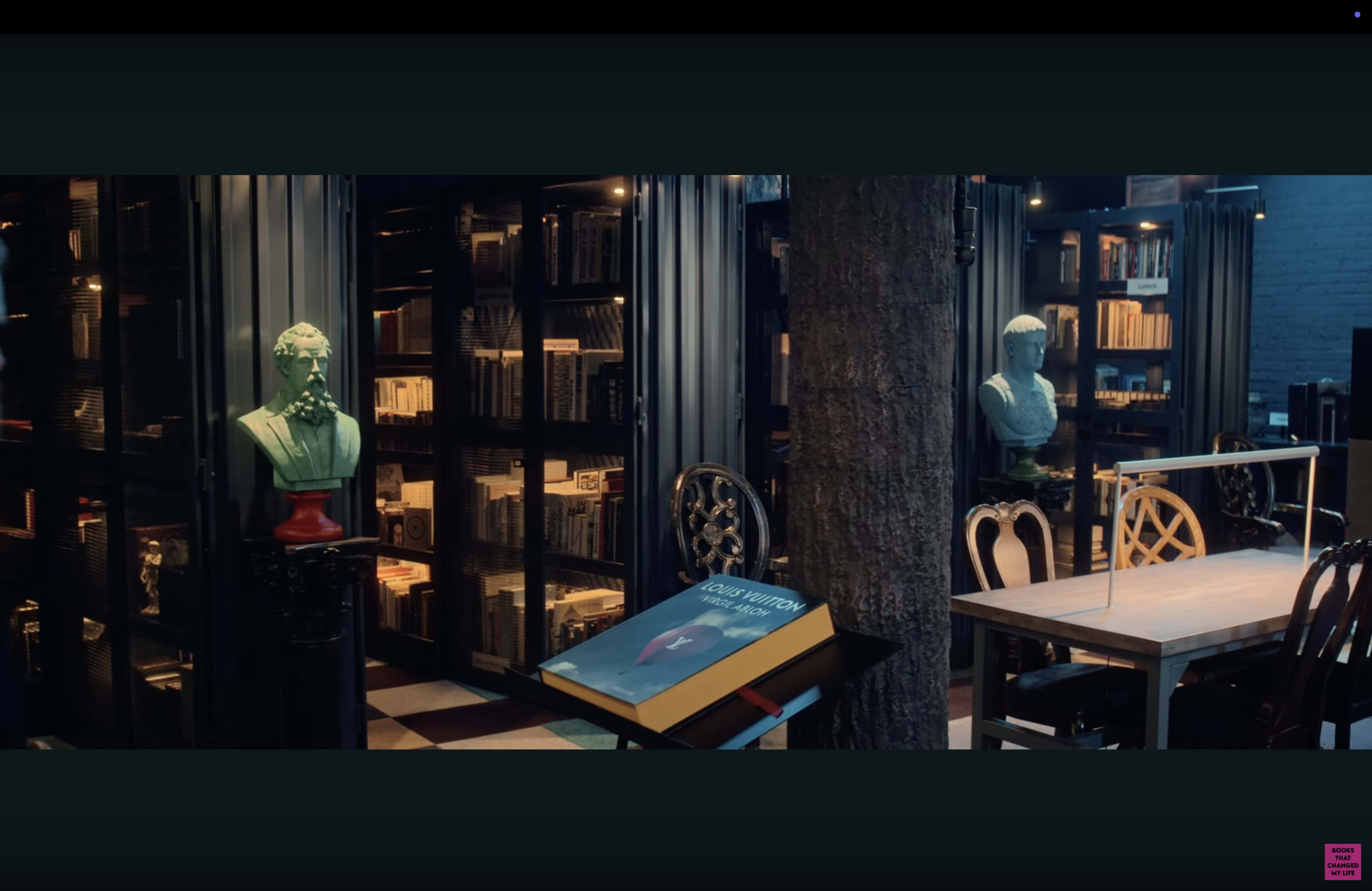 The image shows a stylish library with bookshelves filled with books, a large book on display, and classical busts positioned in the room.