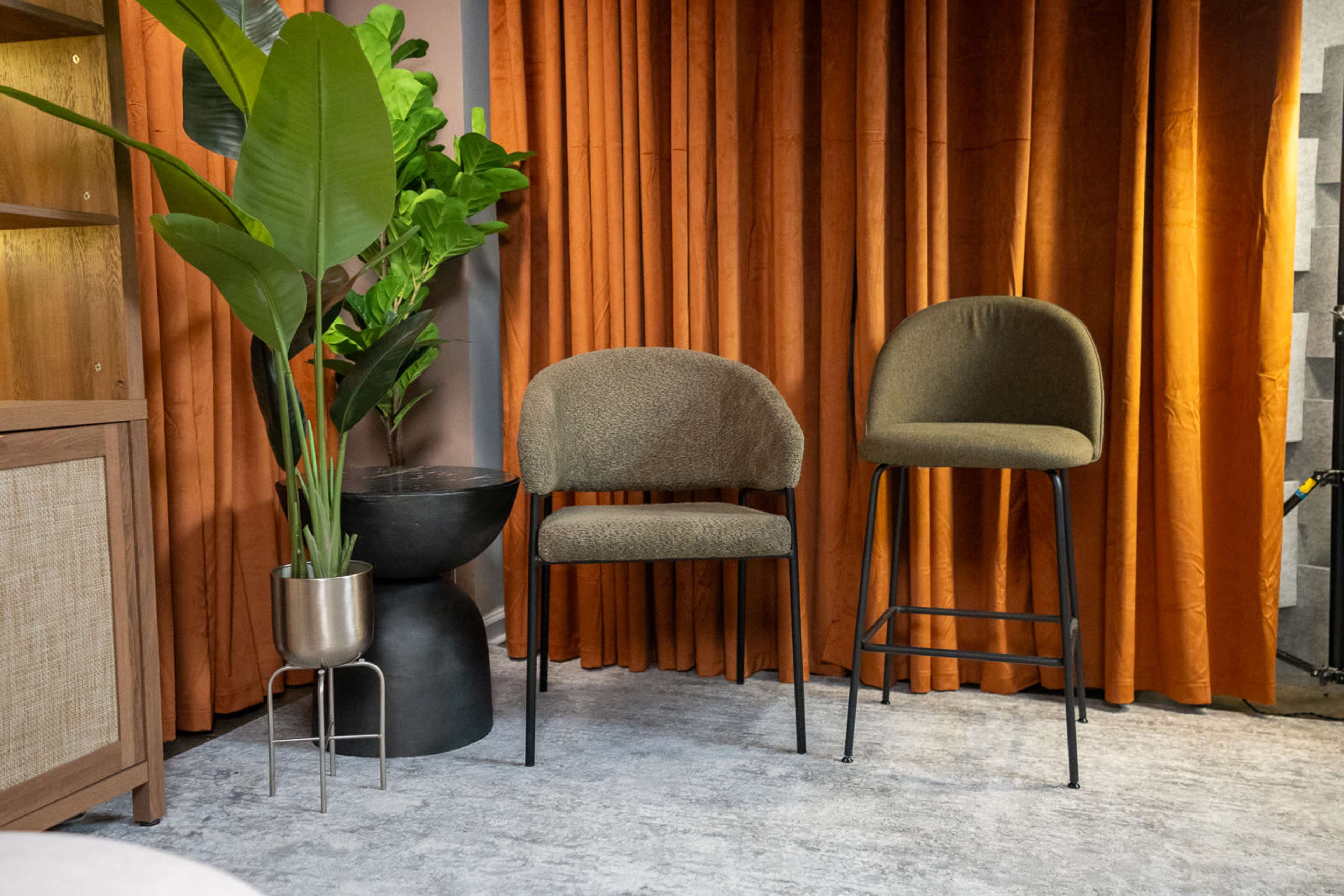 The image shows two chairs, one a dining chair and the other a bar stool, positioned near a potted plant beside a wooden shelf against a backdrop of orange curtains.