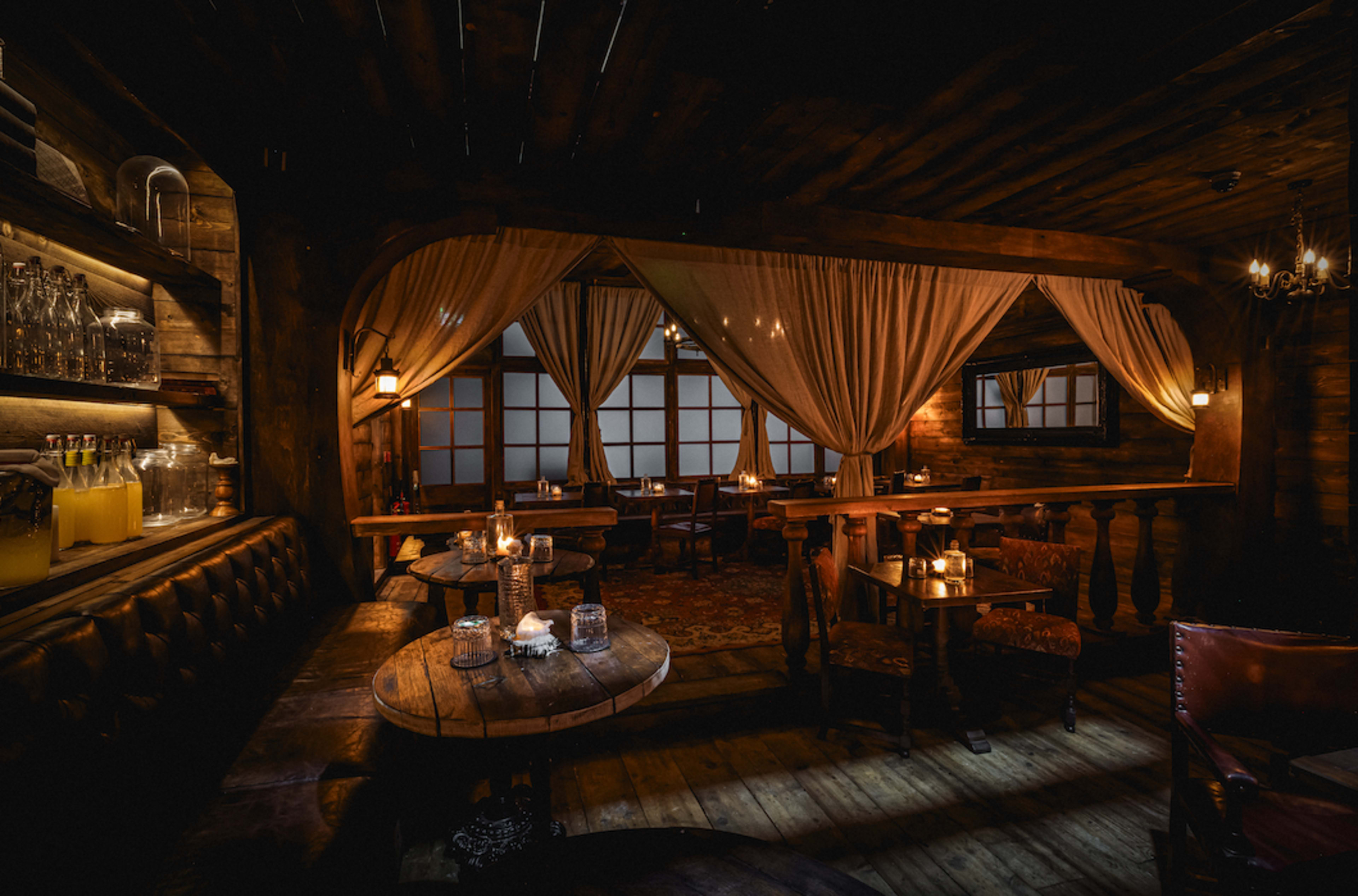 The image shows a dimly lit restaurant interior featuring wooden furnishings, tables with candles, and draped curtains.