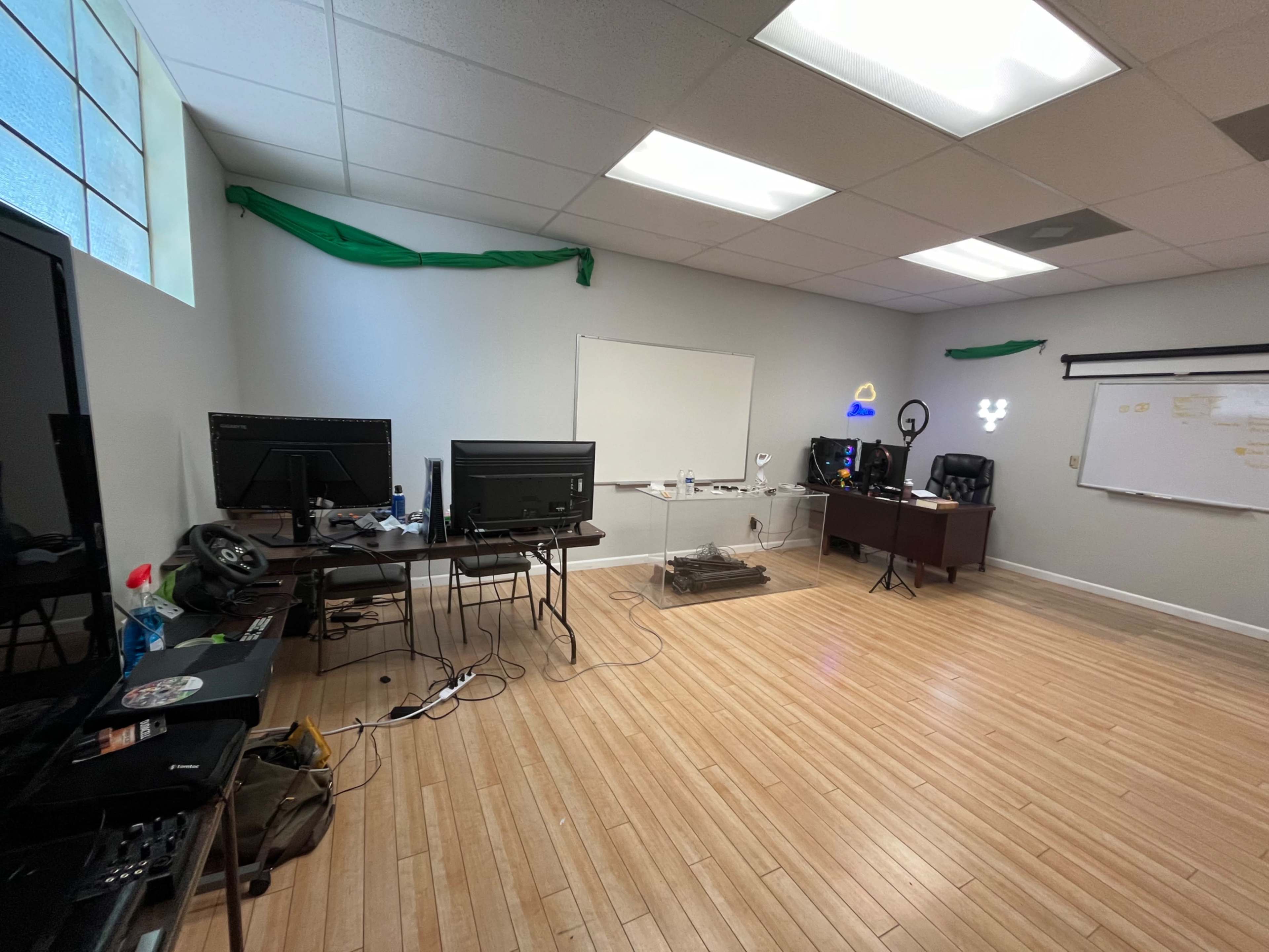 The image shows an office space with wooden floors, two computer monitors on a desk, a whiteboard on the wall, and a small light setup.
