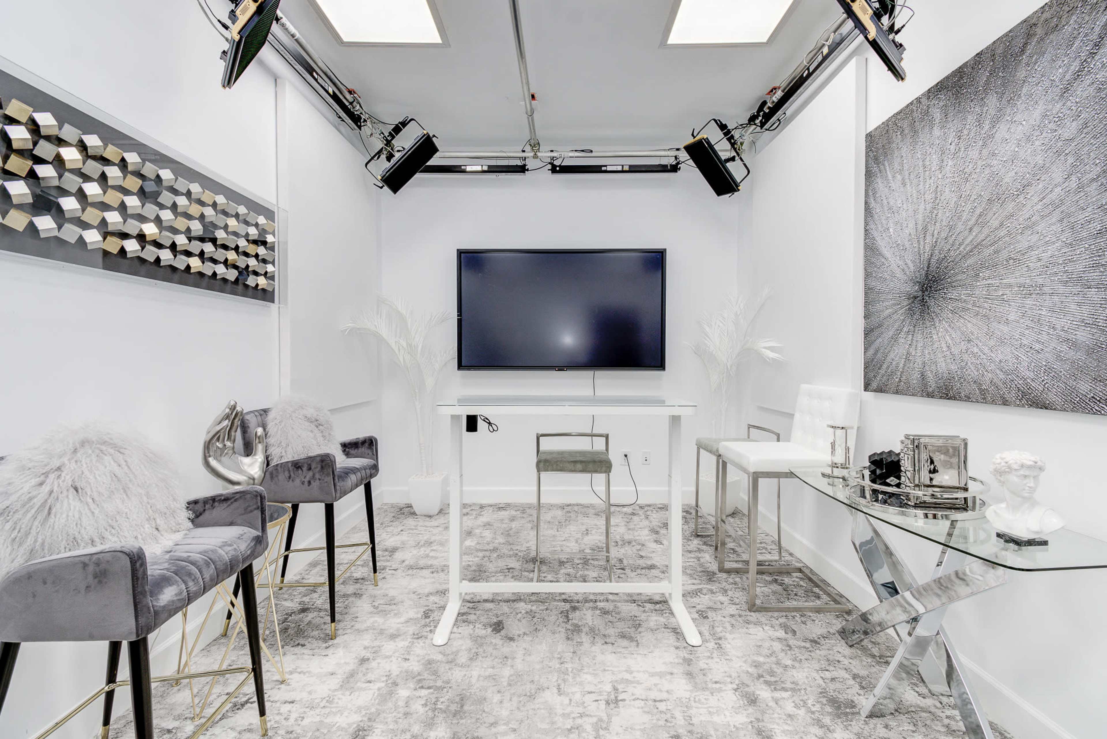 The image shows a modern, minimalist studio with a large television screen, two glass tables, plush seating, and studio lights overhead.
