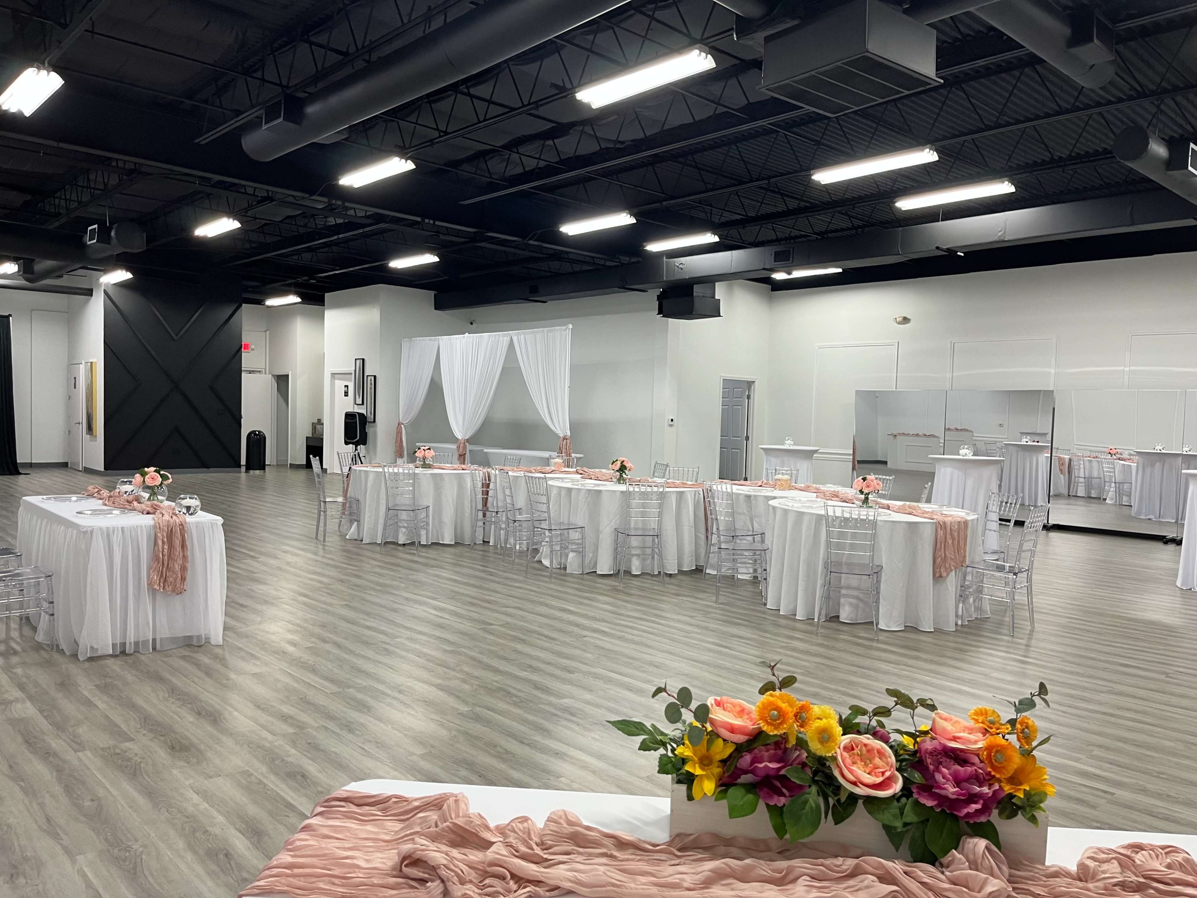 The image shows a spacious event hall with several round tables covered in white tablecloths, adorned with floral centerpieces and draped fabric decorations.