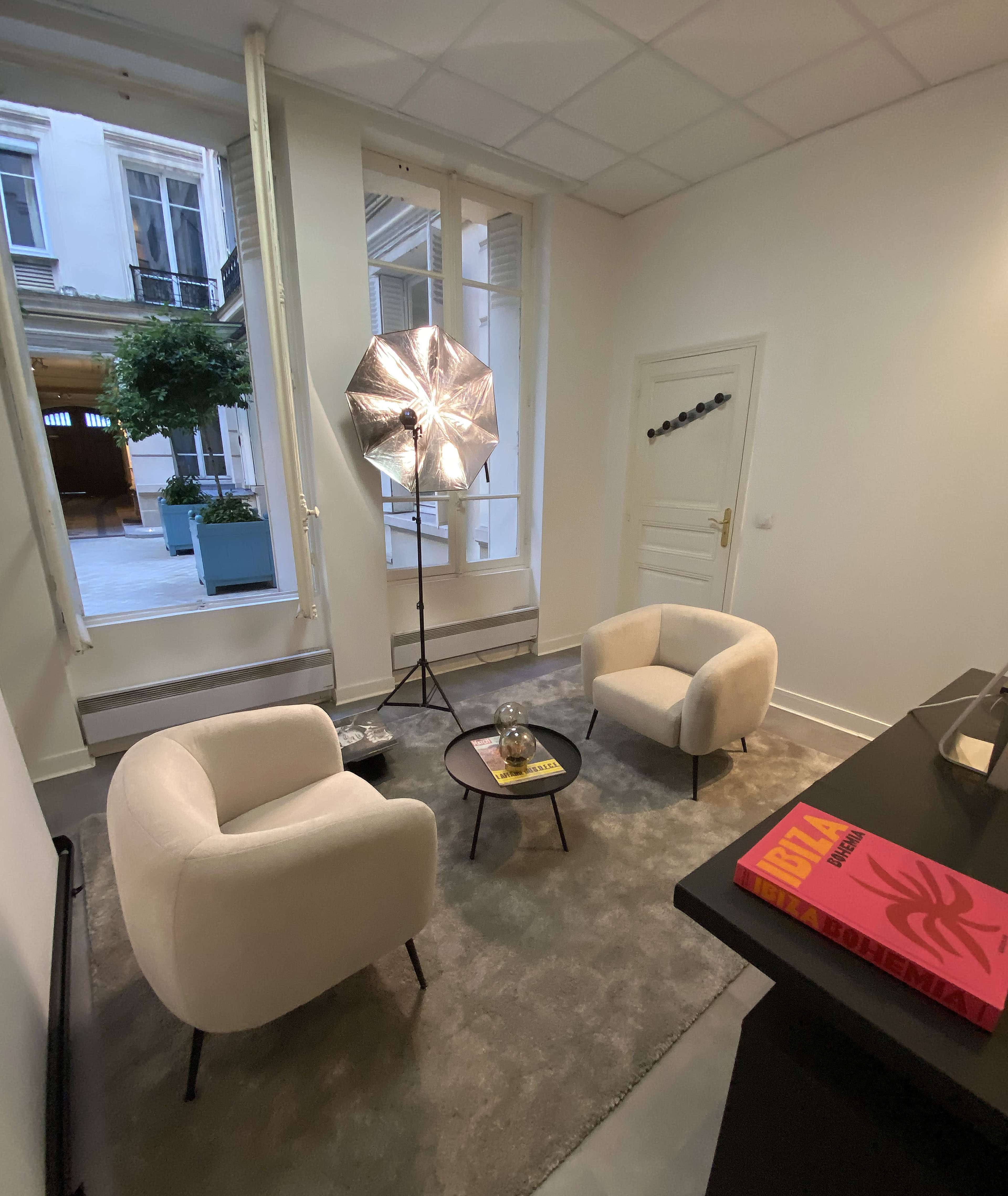 The image shows a small room with two white armchairs facing a black coffee table, a large silver umbrella light, and a window overlooking a courtyard with potted plants.