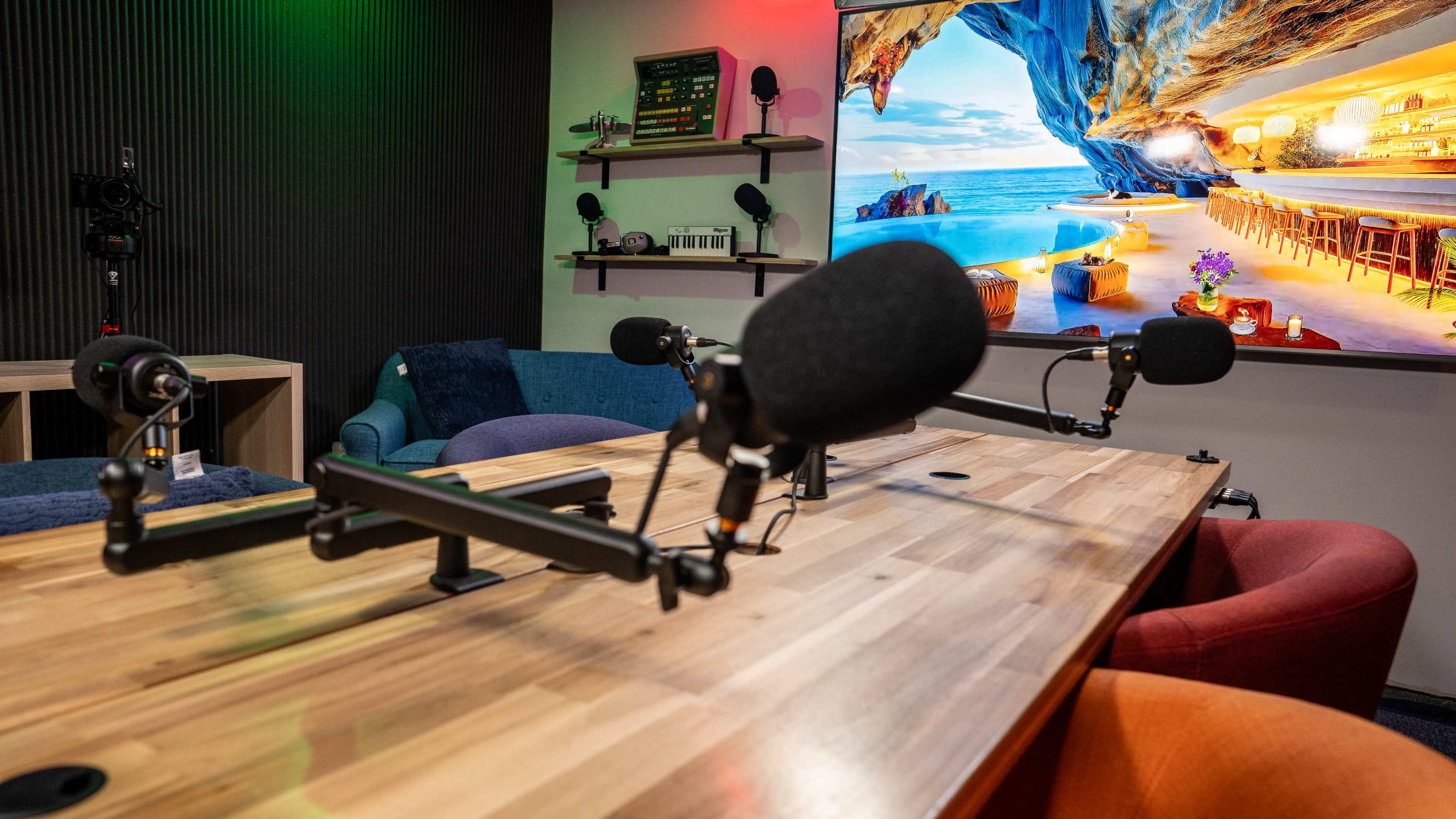A podcast recording studio features a wooden table with microphone stands and colorful chairs, set against a large scenic backdrop.