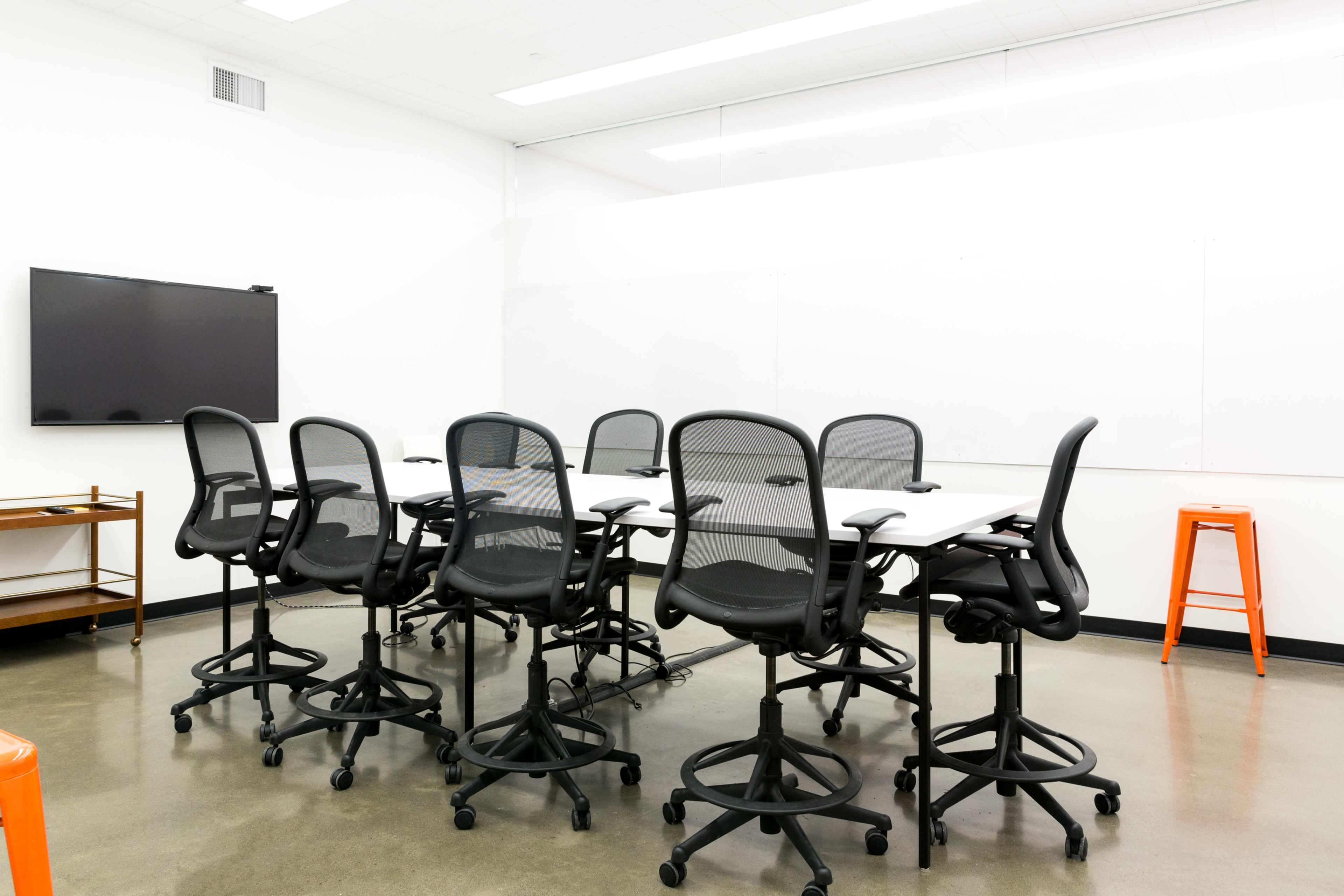 A conference room features a long table surrounded by adjustable chairs, with a television mounted on the wall and two orange stools in the corner.