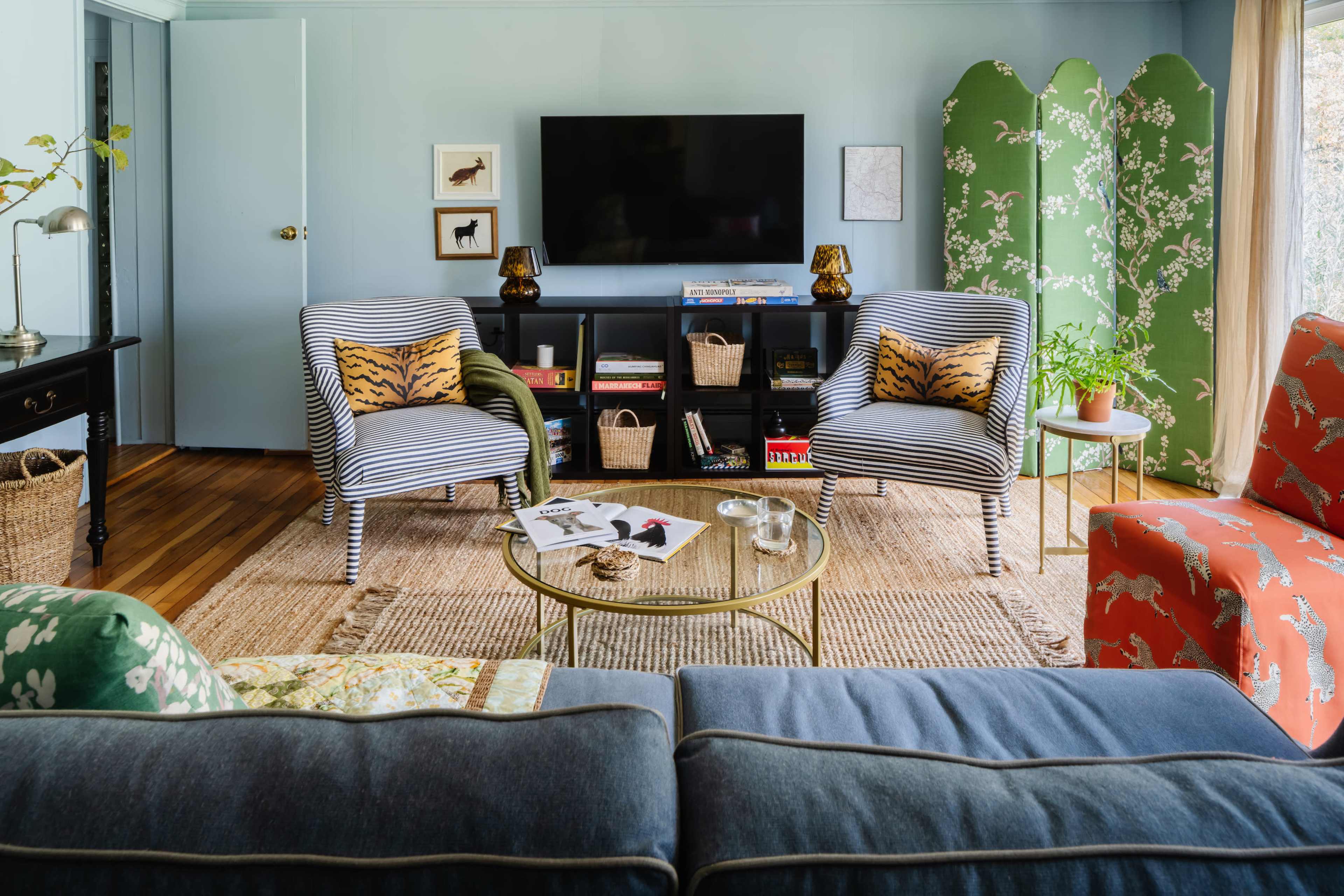 A living room features two striped armchairs with animal-patterned cushions, a glass coffee table, and a television mounted on a wall, surrounded by decorative plants and woven decor.