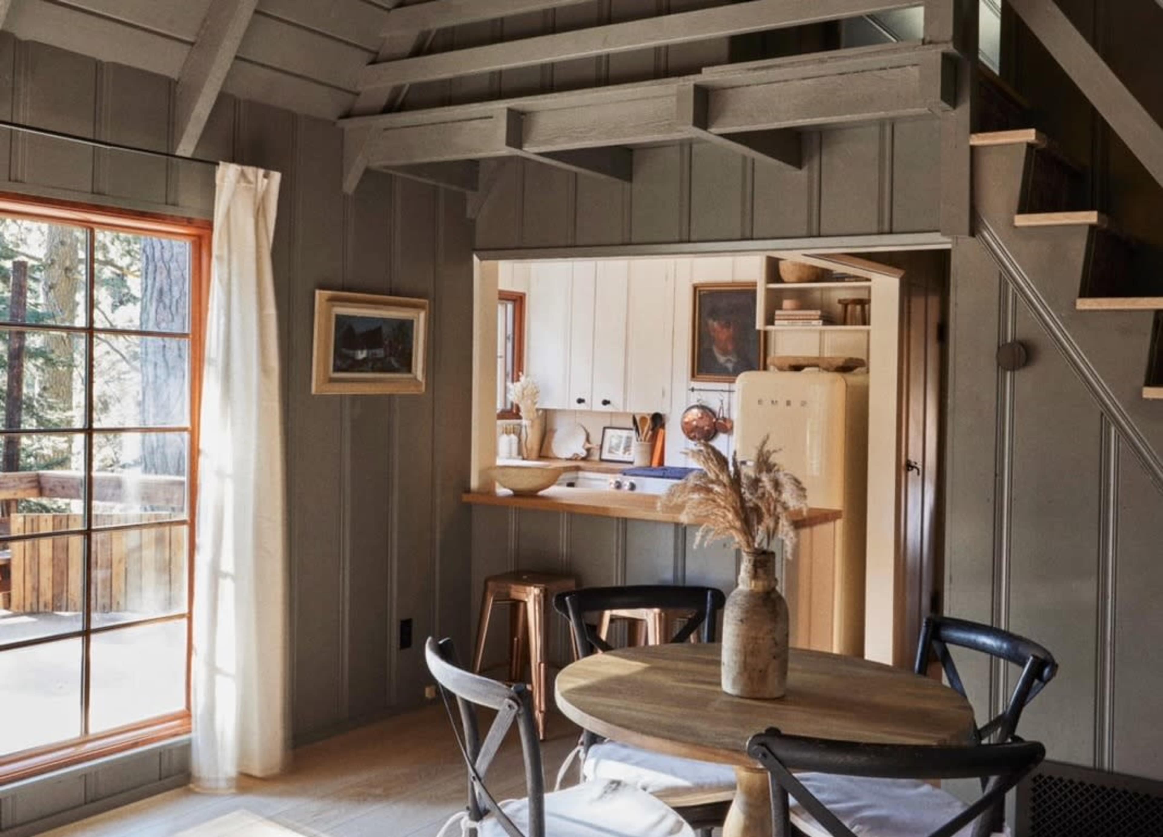 A cozy, rustic interior of a cabin with a round wooden dining table, a vintage fridge, and a staircase leading to an upper level.