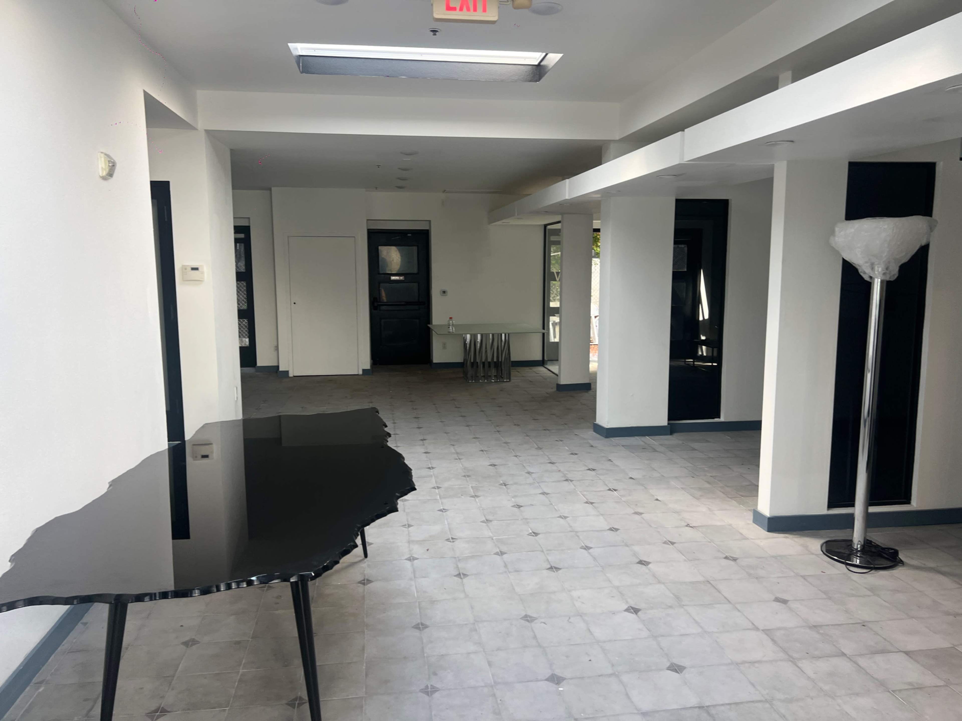 The image shows a spacious, empty room with a black table on the left and a floor lamp on the right, featuring tile flooring and white walls.