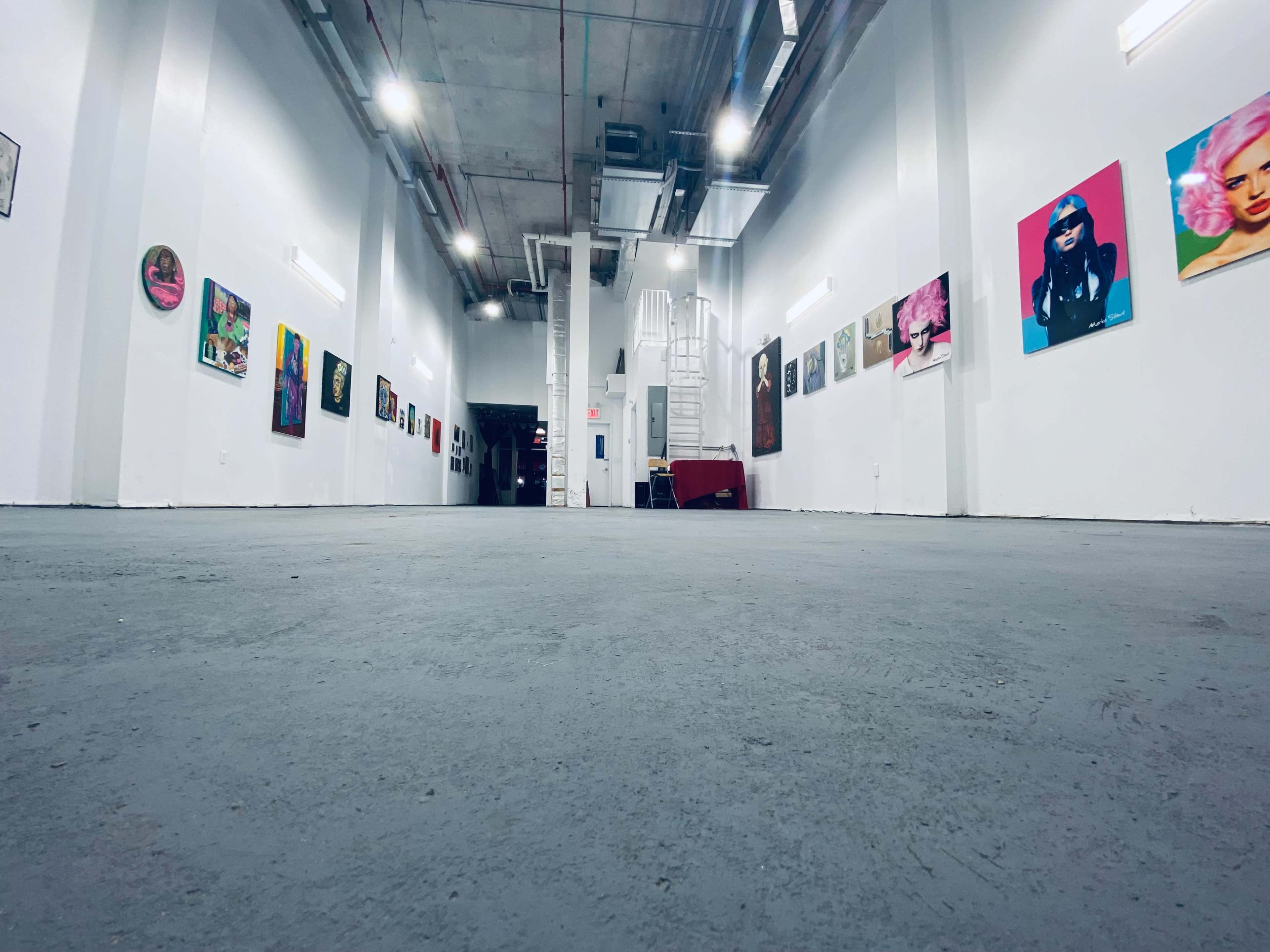 An art gallery with white walls adorned with various colorful paintings, and a staircase visible at the back.