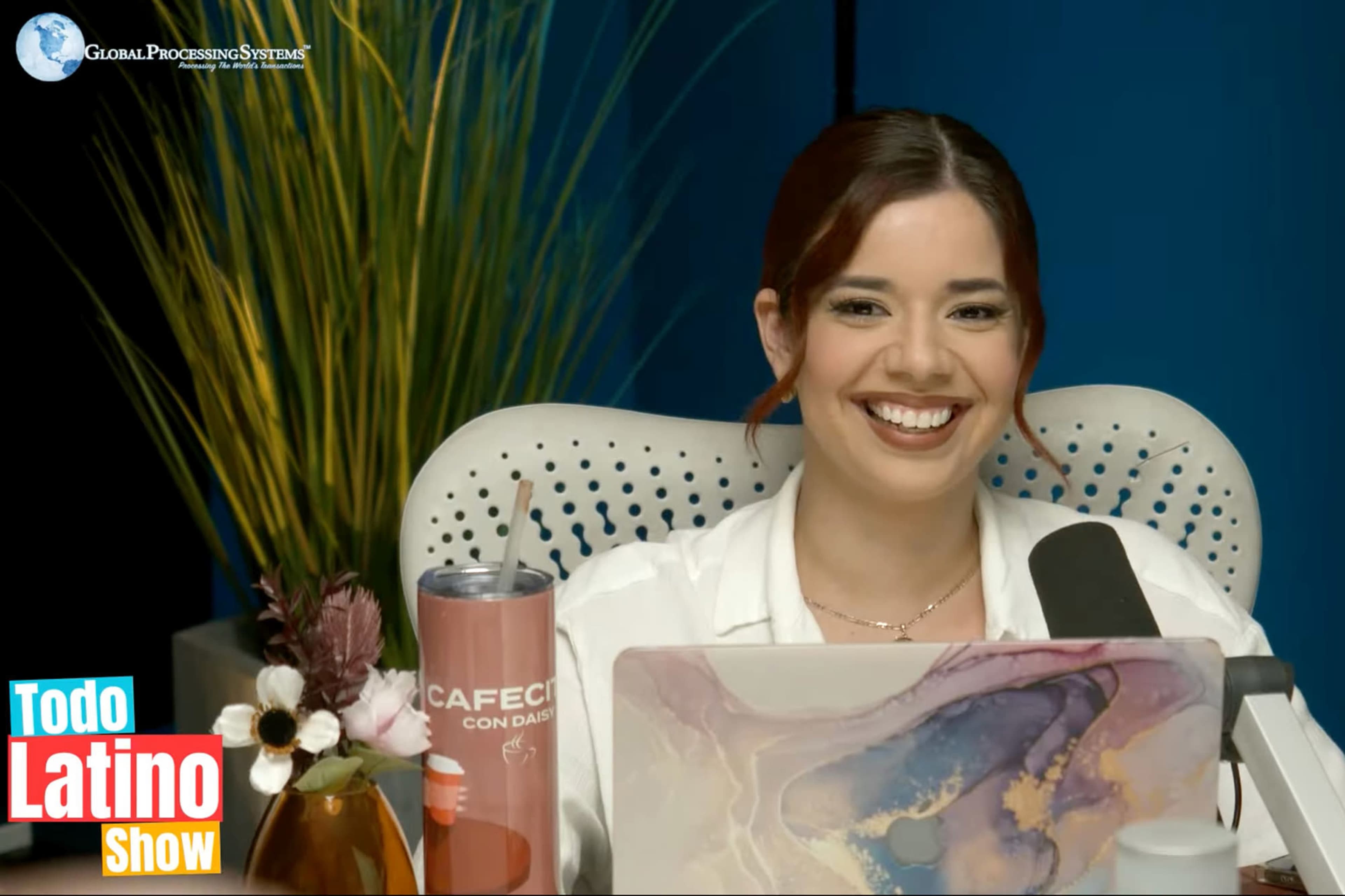 A woman is sitting at a desk with a colorful laptop, smiling at the camera, surrounded by decorative plants and a drink.