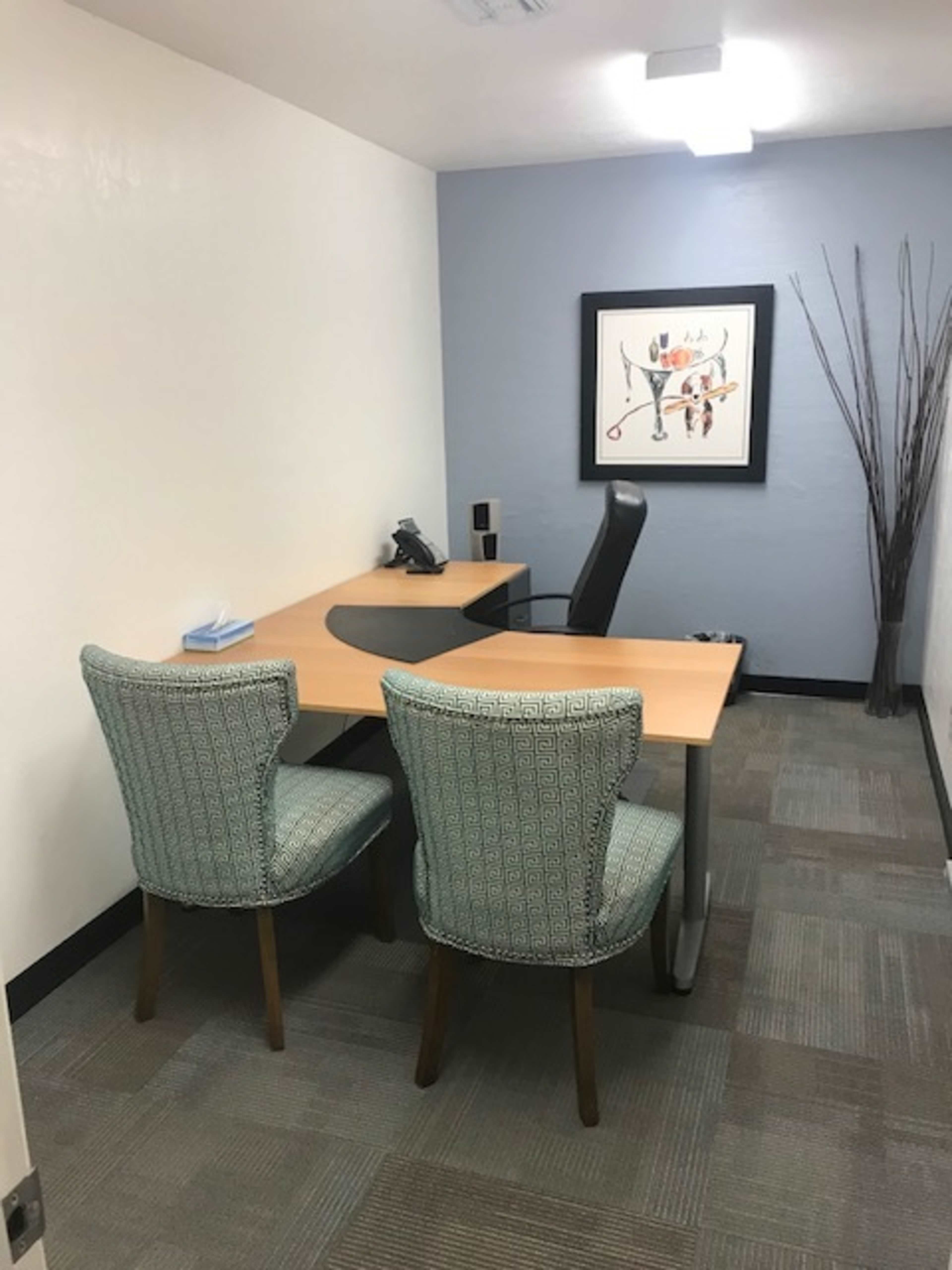 The image shows a small office space with a wooden desk, an office chair, two patterned chairs, and a piece of artwork on the wall.