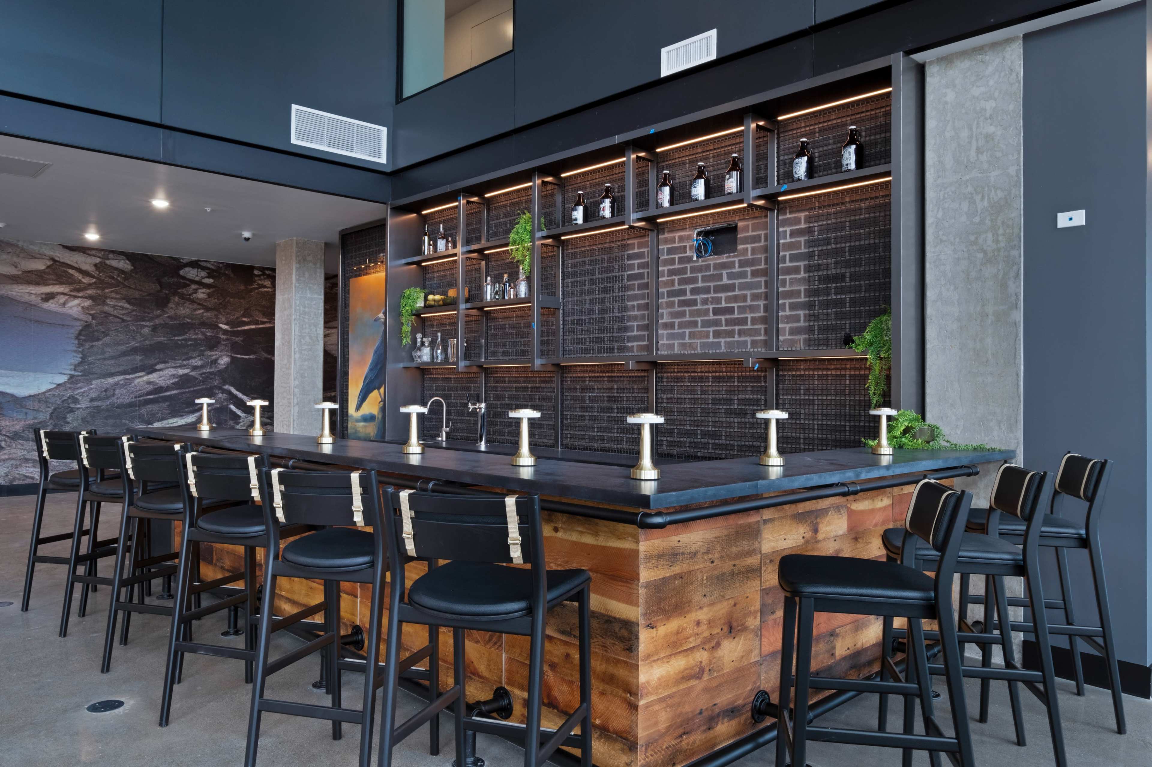 The image shows a modern bar with a wooden counter, black chairs, and a wall featuring shelves that hold bottles against a brick backdrop.
