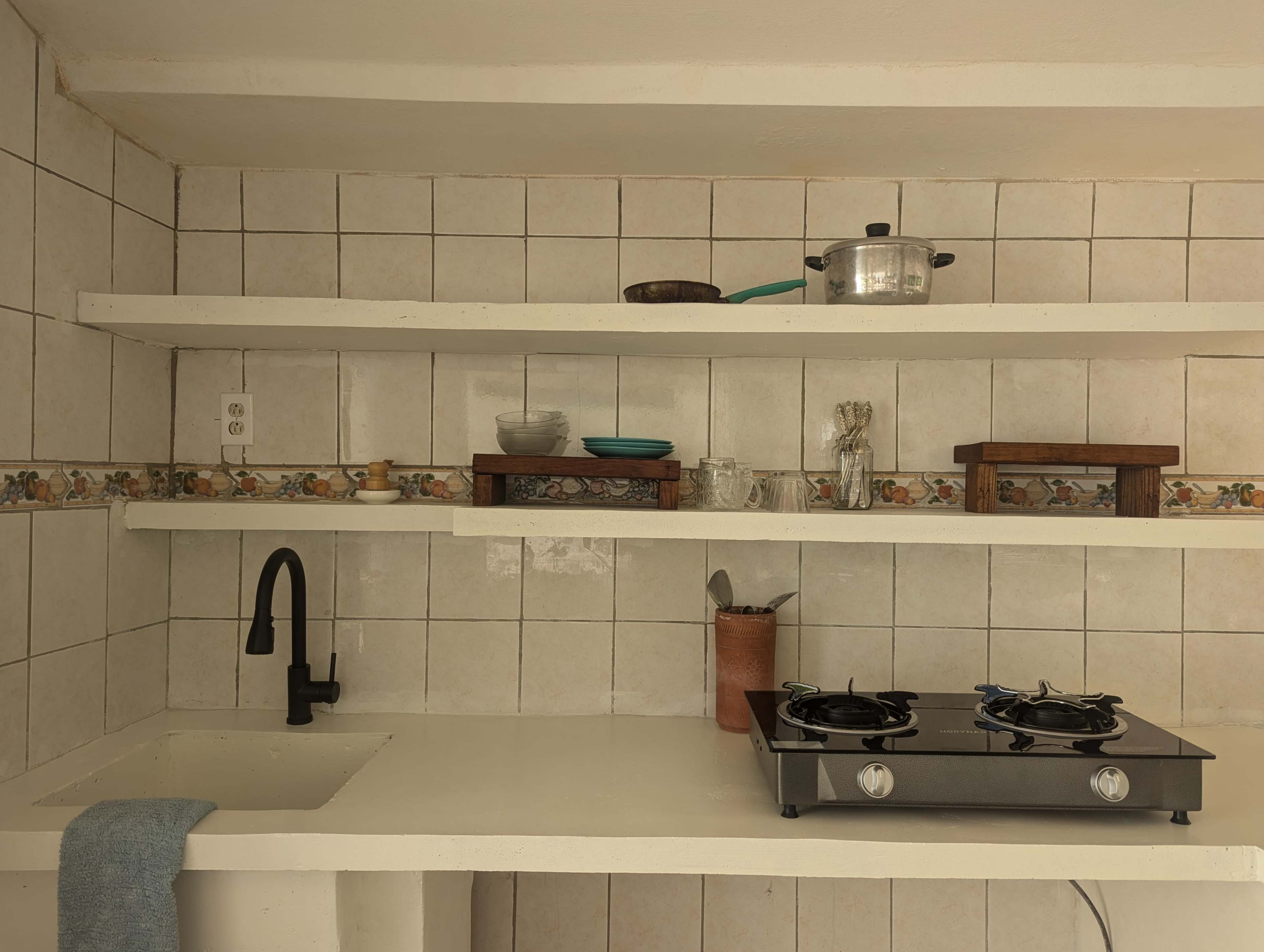 The image shows a simple kitchen with white tiled walls, a gas stove, a sink, and several shelves holding cooking utensils and items.