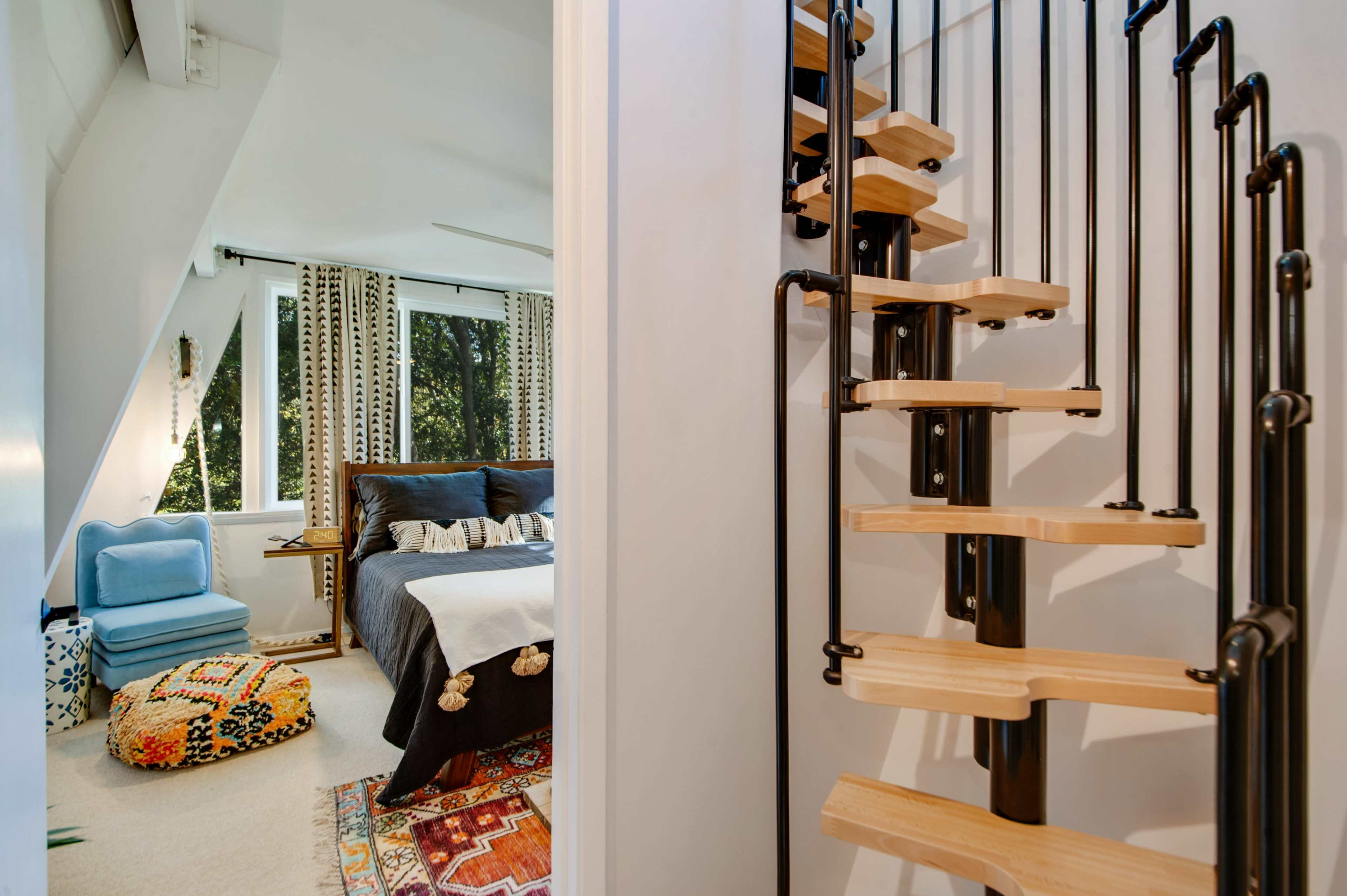A spiral staircase with wooden steps leads up to a bedroom featuring a bed, decorative pillows, and large windows with patterned curtains.