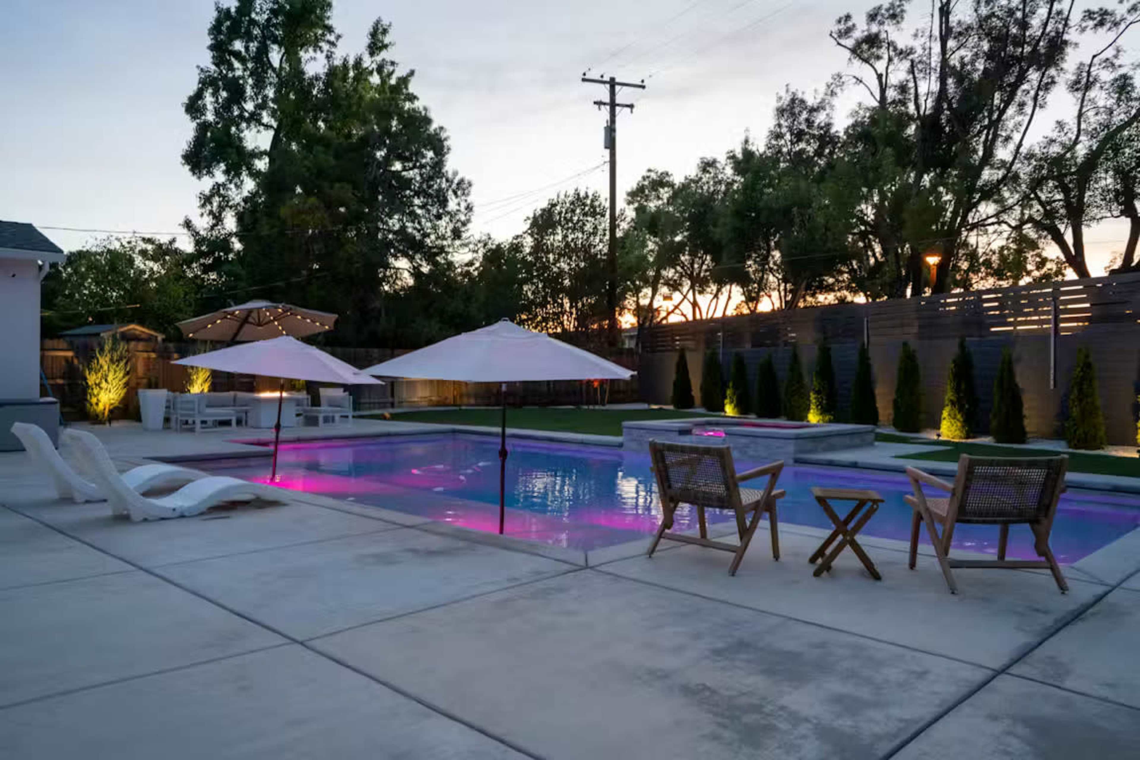The image shows a backyard pool area with lounge chairs, two umbrellas, and a seating arrangement, illuminated by colored lights as the sun sets.