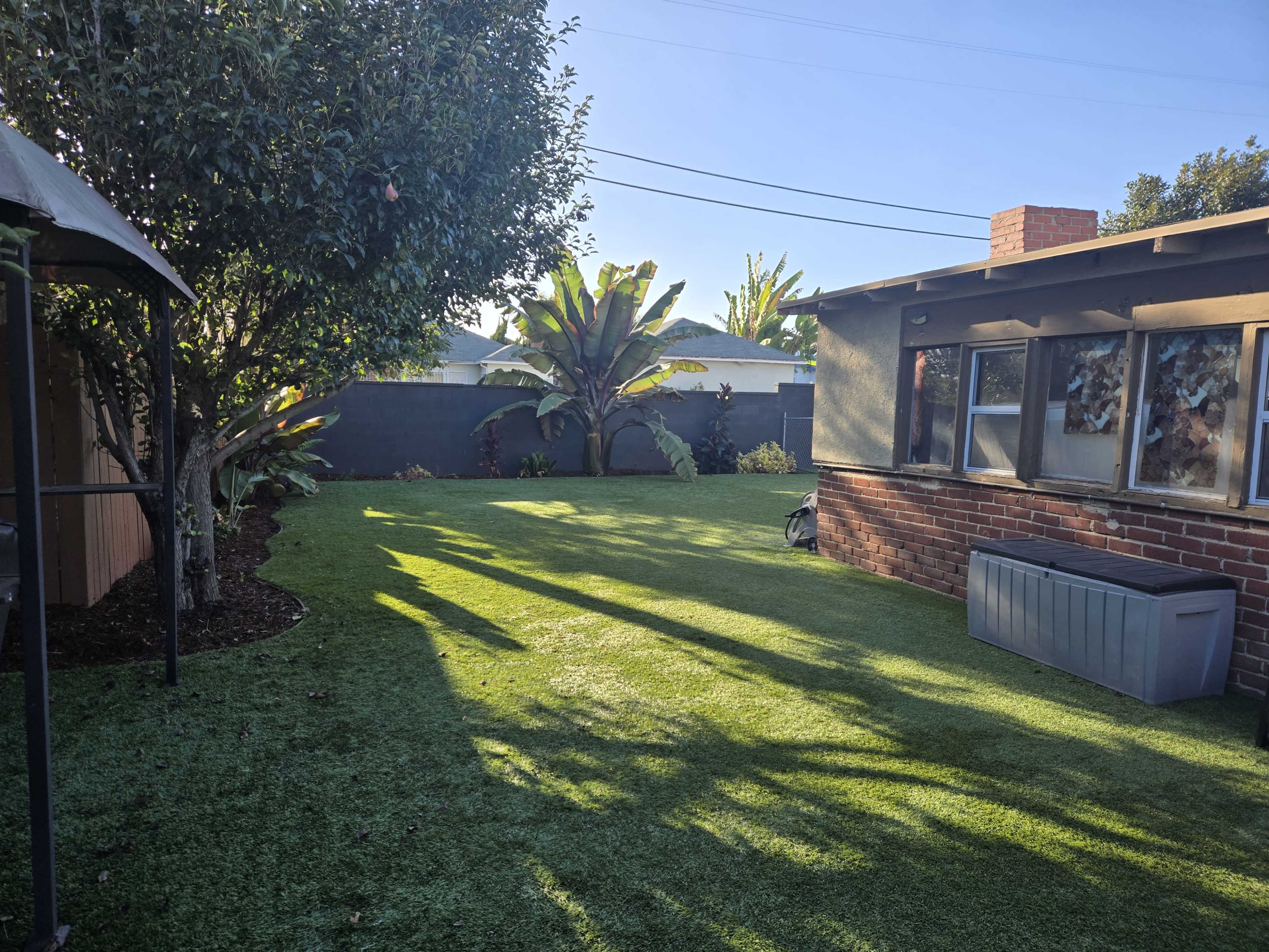 The image shows a well-maintained backyard with green artificial turf, bordered by trees and plants, alongside a brick house.