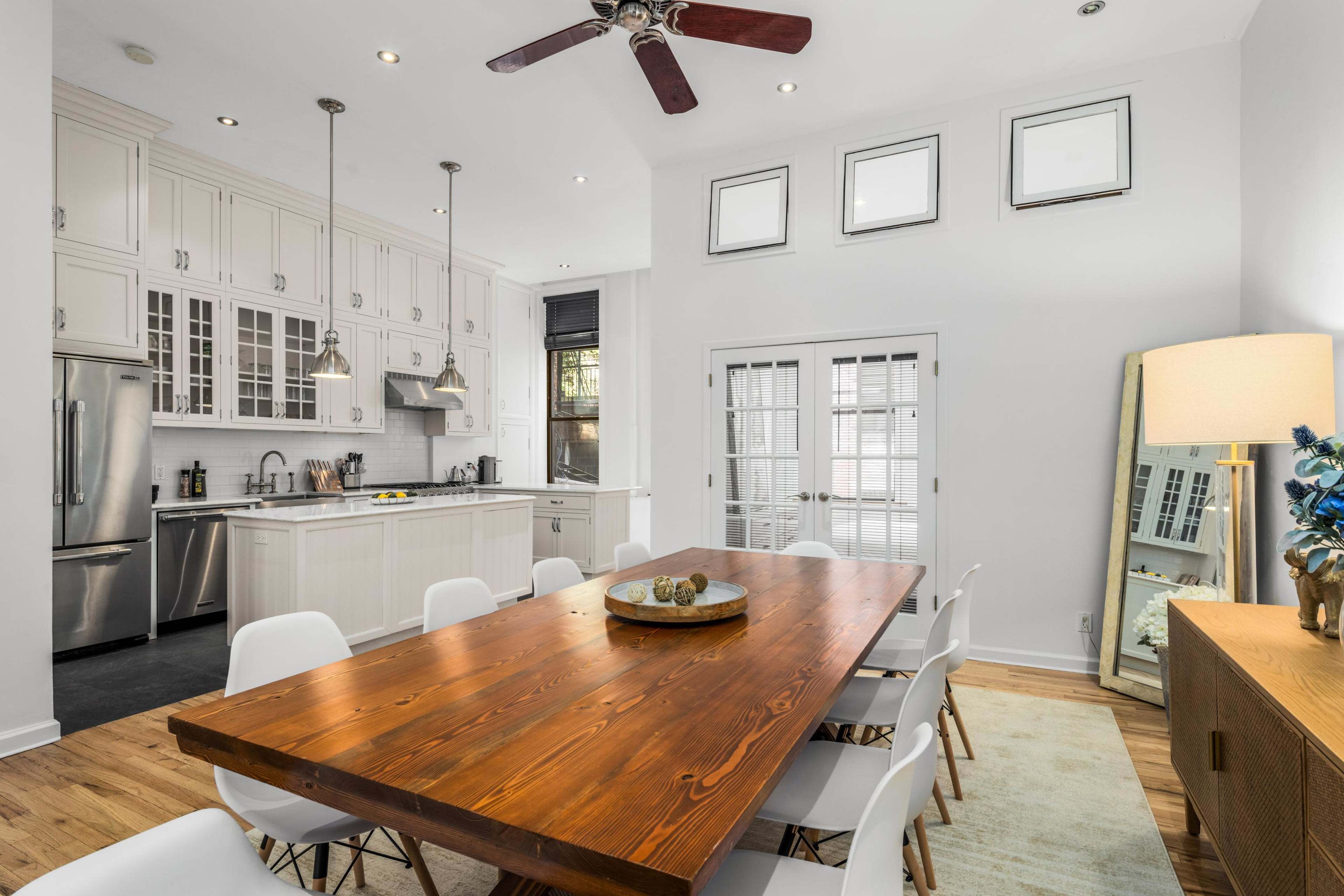 A modern kitchen and dining area featuring a large wooden dining table surrounded by white chairs, with a sleek kitchen equipped with stainless steel appliances and white cabinetry.