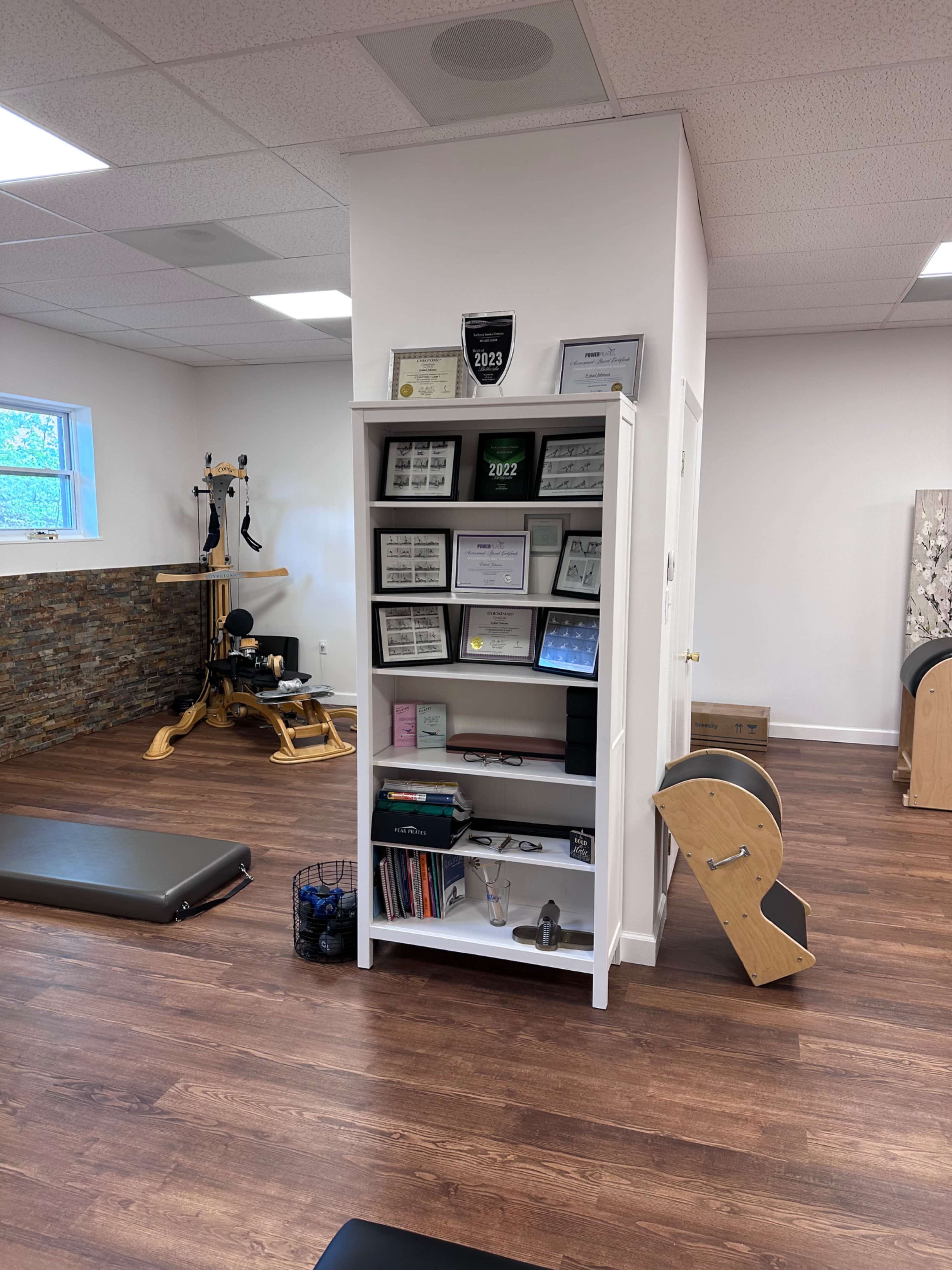 The image shows a fitness studio with a white shelving unit displaying various certificates and materials, a reformer machine in the background, and exercise mats on the floor.