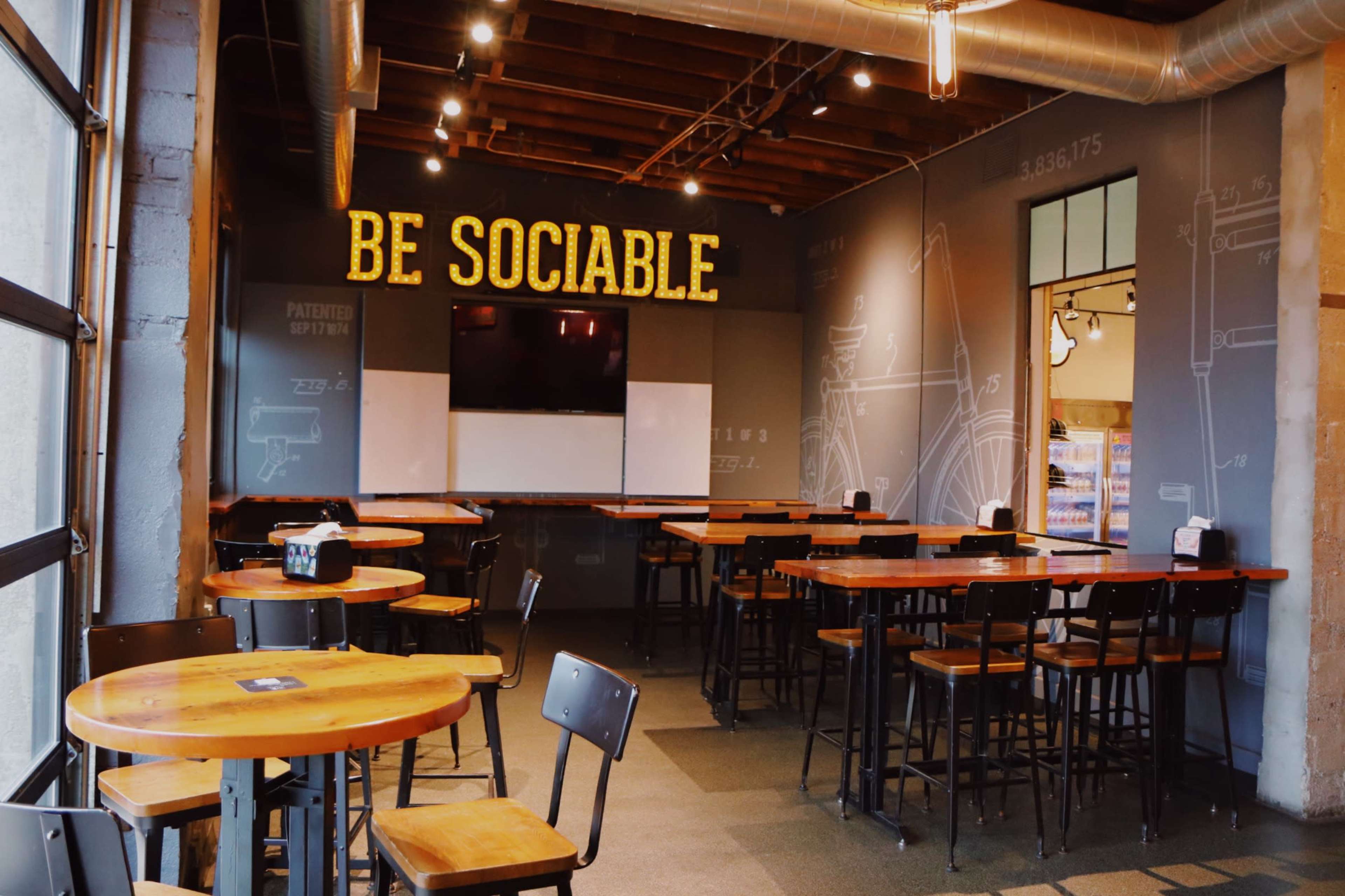 A restaurant interior with wooden tables and chairs, featuring a wall with the phrase "BE SOCIABLE" illuminated above a TV screen and whiteboard.