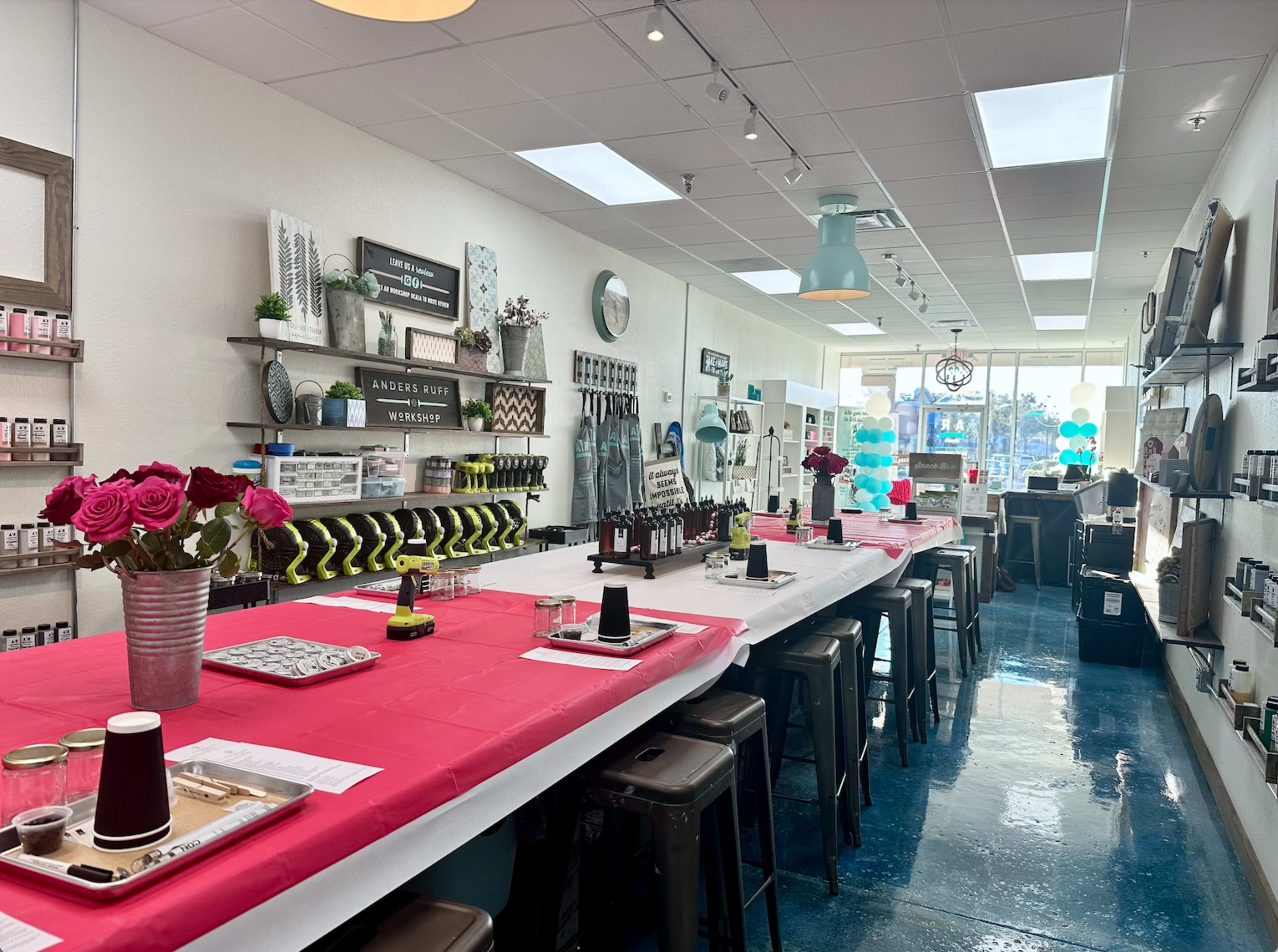 A brightly lit craft studio features long tables set with pink tablecloths, decorative flowers, art supplies, and various project materials along the walls.