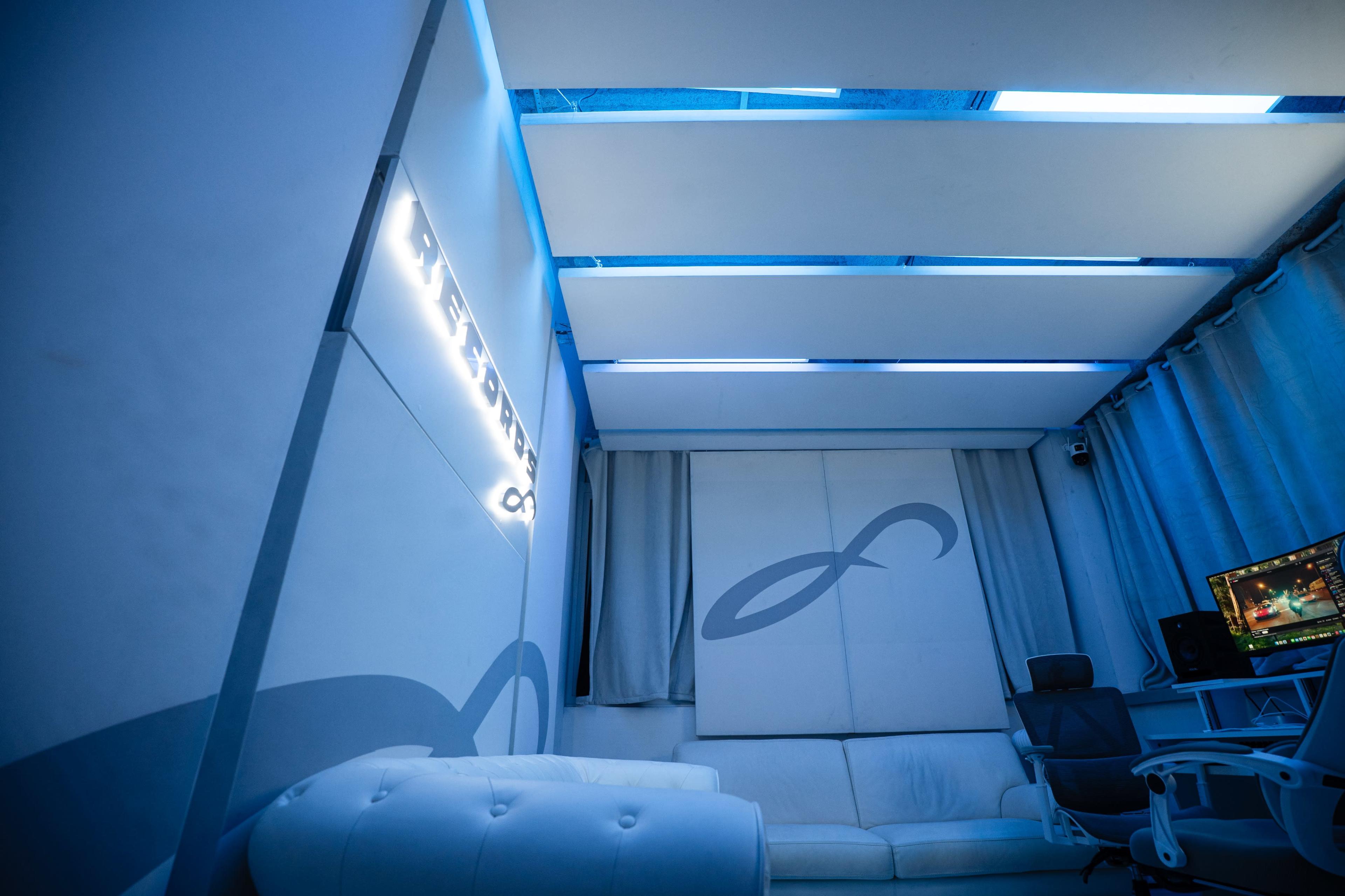The image shows a modern, minimalistic room with blue lighting, a white sofa, and a computer desk with a monitor.