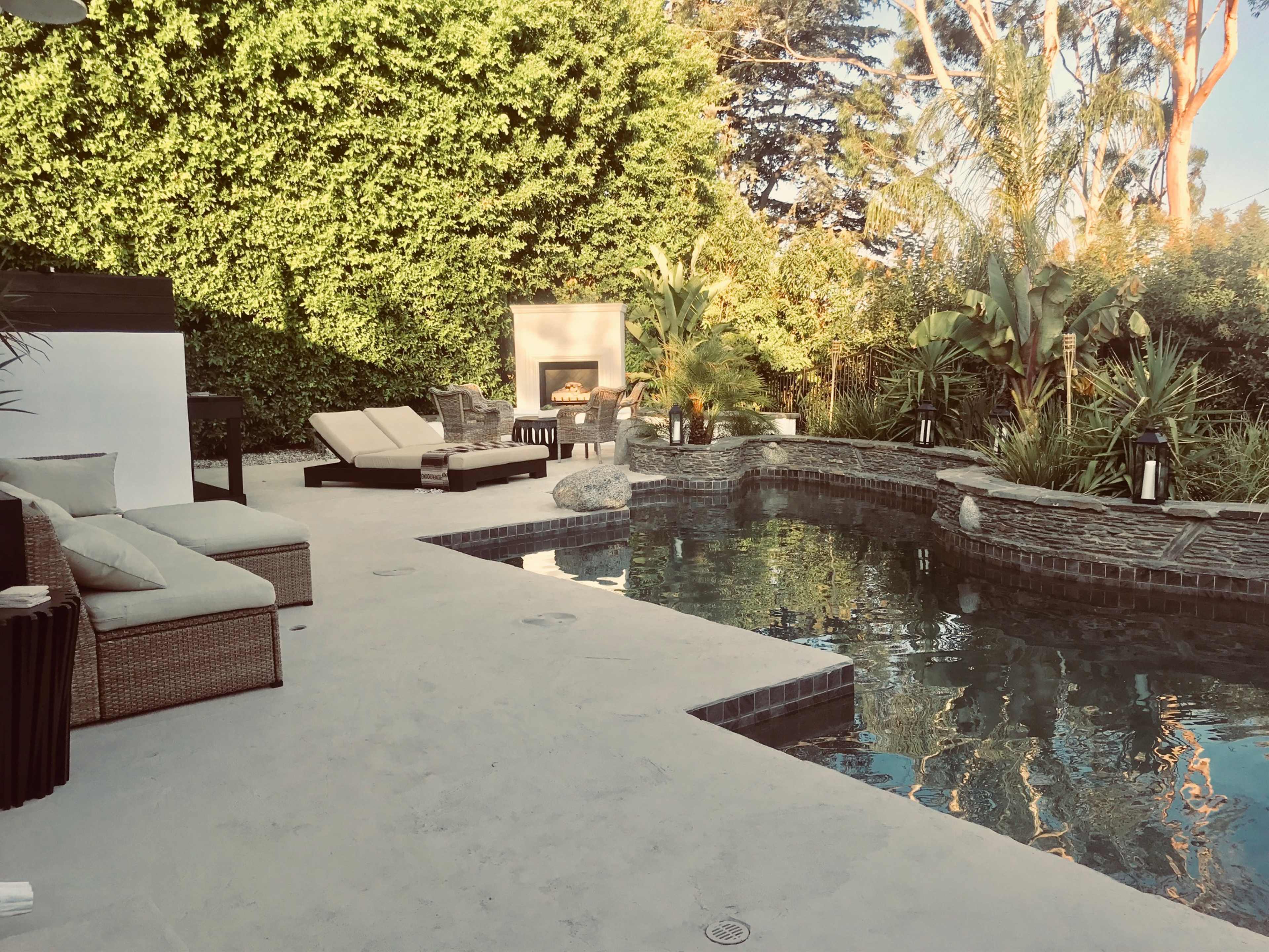 The image shows a backyard pool area with lounge chairs, a fire pit, and lush greenery surrounding the water.