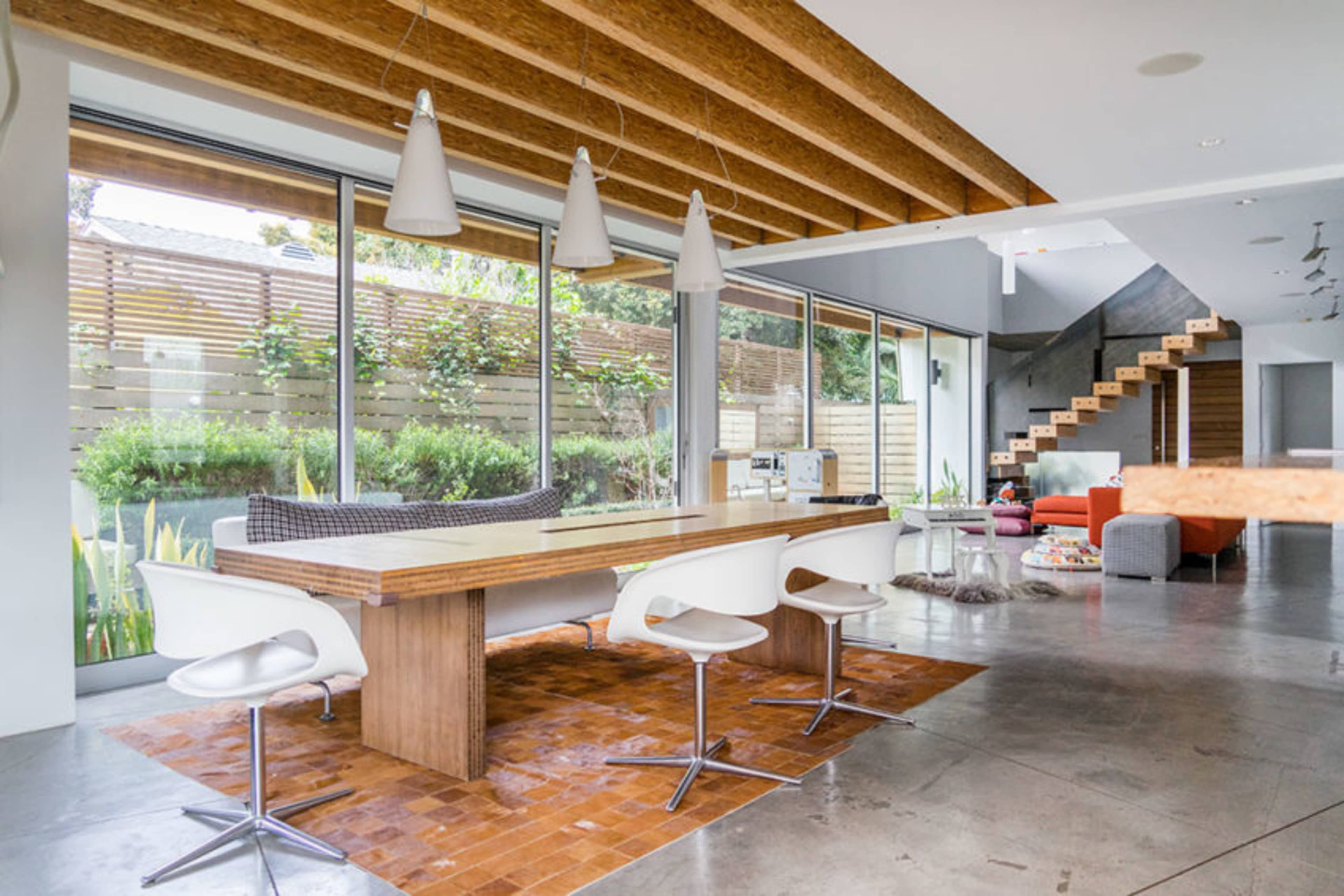 The image shows a modern living space with a large wooden dining table, sleek white chairs, and large windows overlooking a landscaped outdoor area.