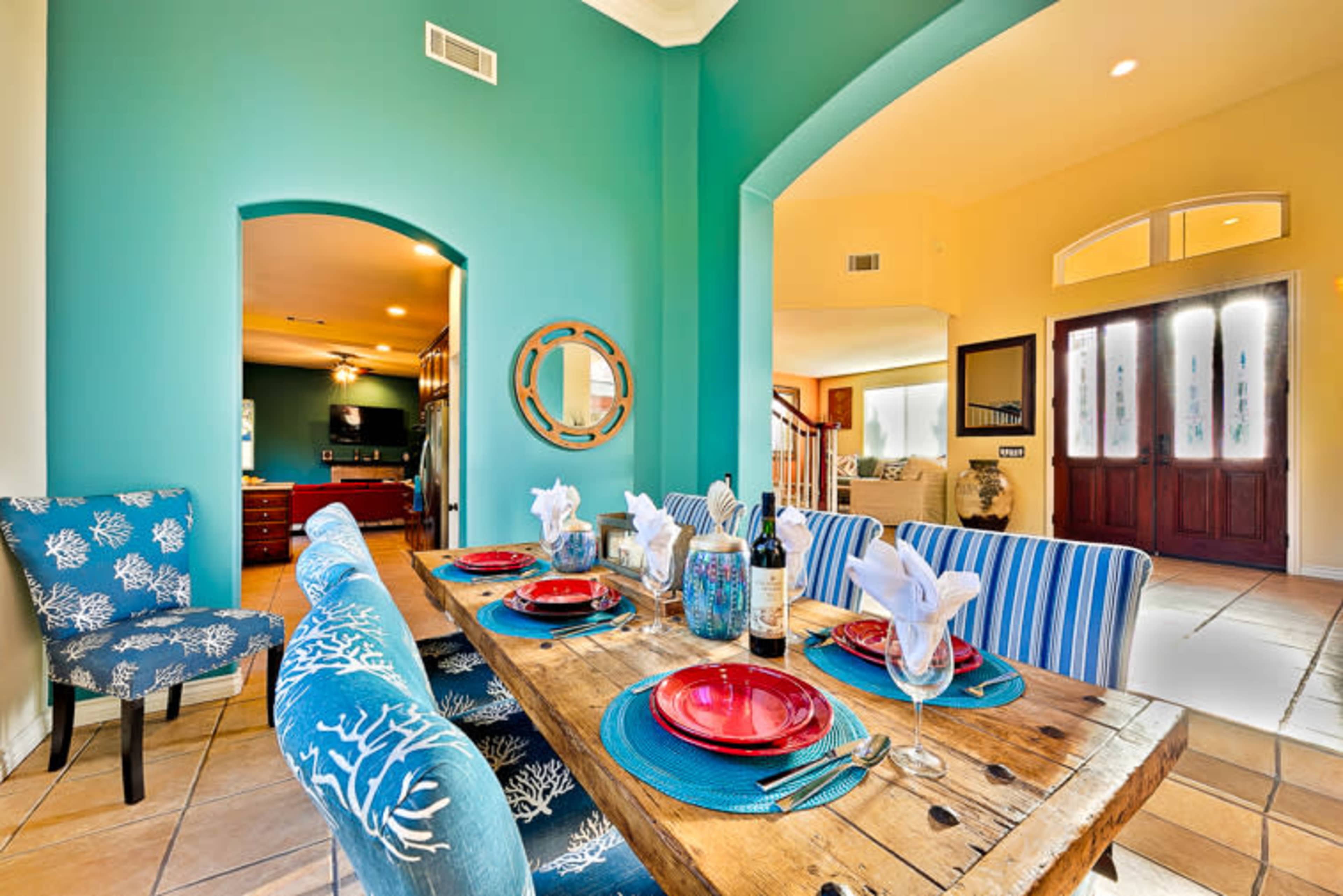 A dining area features a wooden table set with plates and utensils, surrounded by colorful chairs against a turquoise wall and an open entryway.