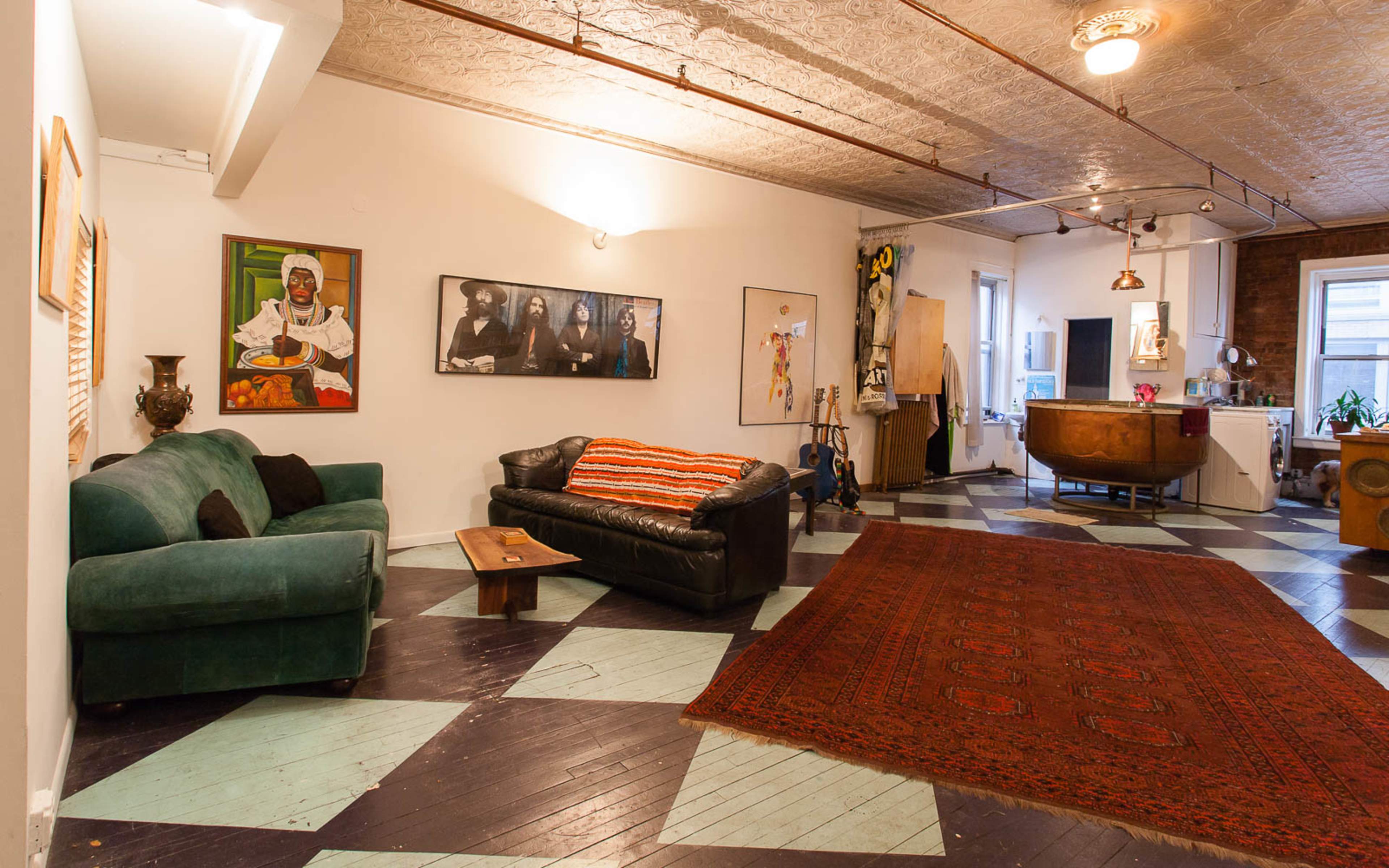 A spacious room featuring a green couch, a black leather sofa with a red-striped throw, a patterned rug on the floor, and various artworks on the walls.