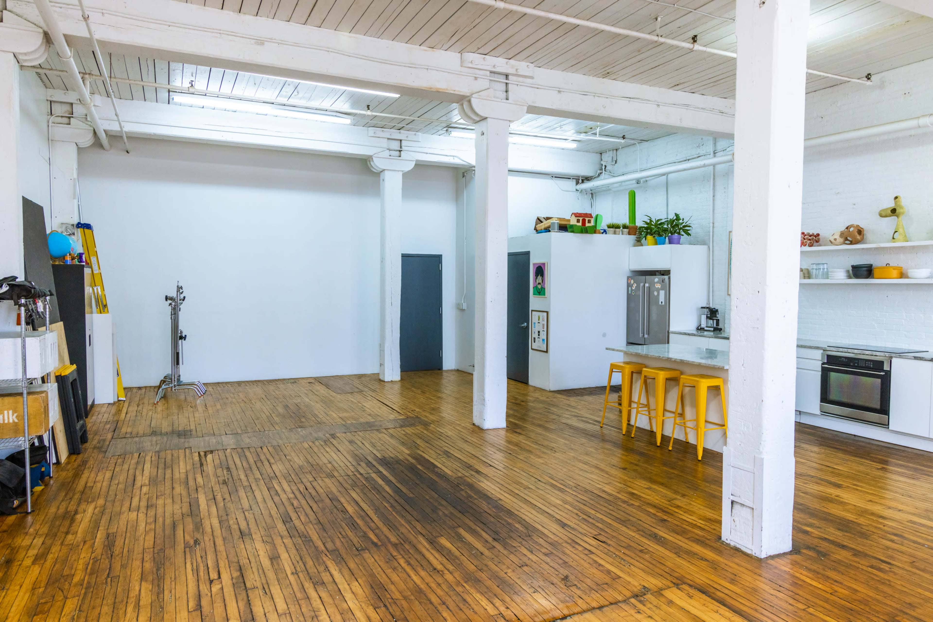 A spacious, minimalist studio with wooden flooring, white walls, and a kitchenette featuring yellow bar stools.