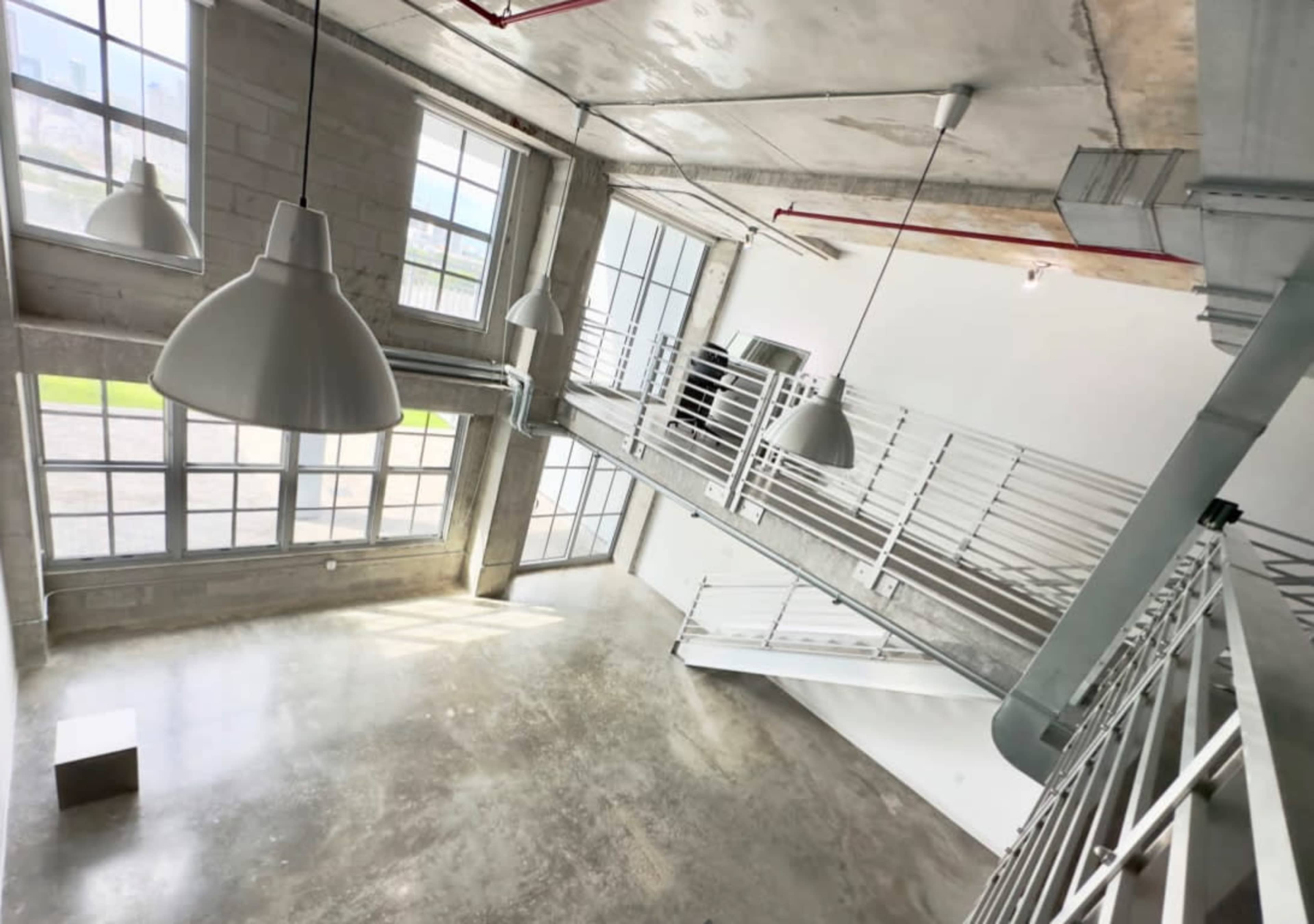 The image shows a modern, spacious interior with large windows, concrete walls, and a multi-level design featuring white railings and pendant lighting.